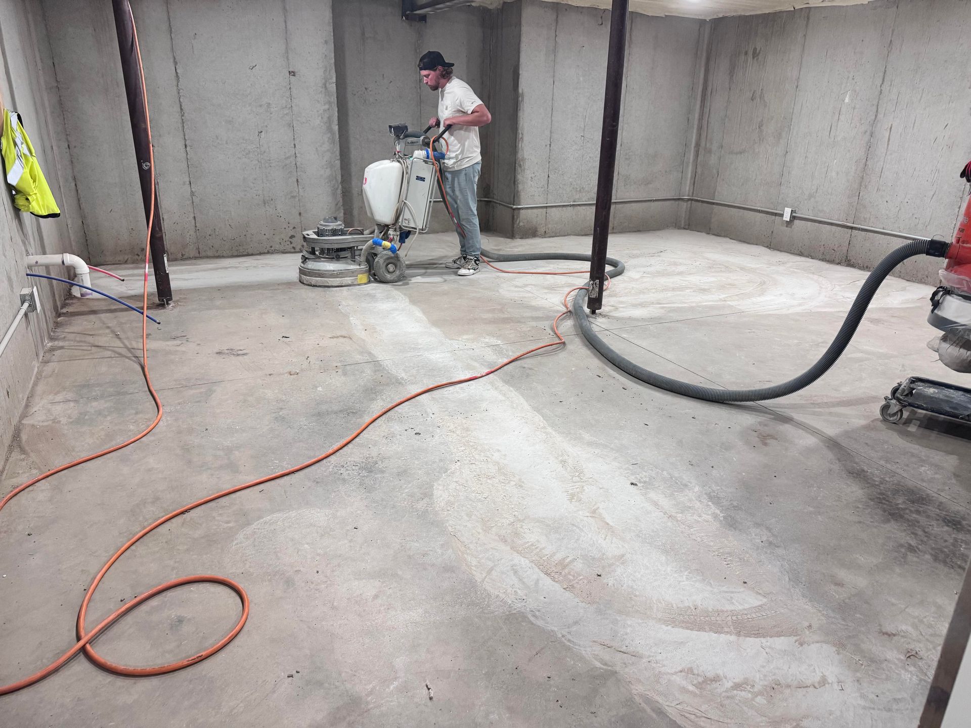 Man grinding concrete floor in a basement. Dusty, with grinding machine, power cords, and walls.