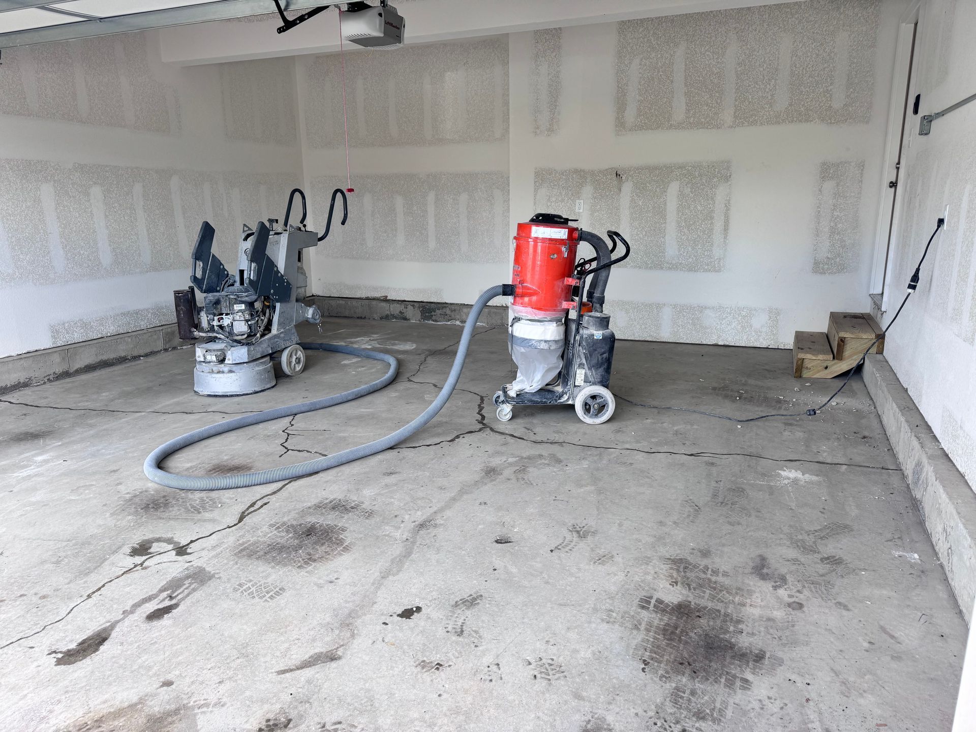 Two concrete grinding machines in a garage, connected by a hose. Gray concrete floor with cracks.