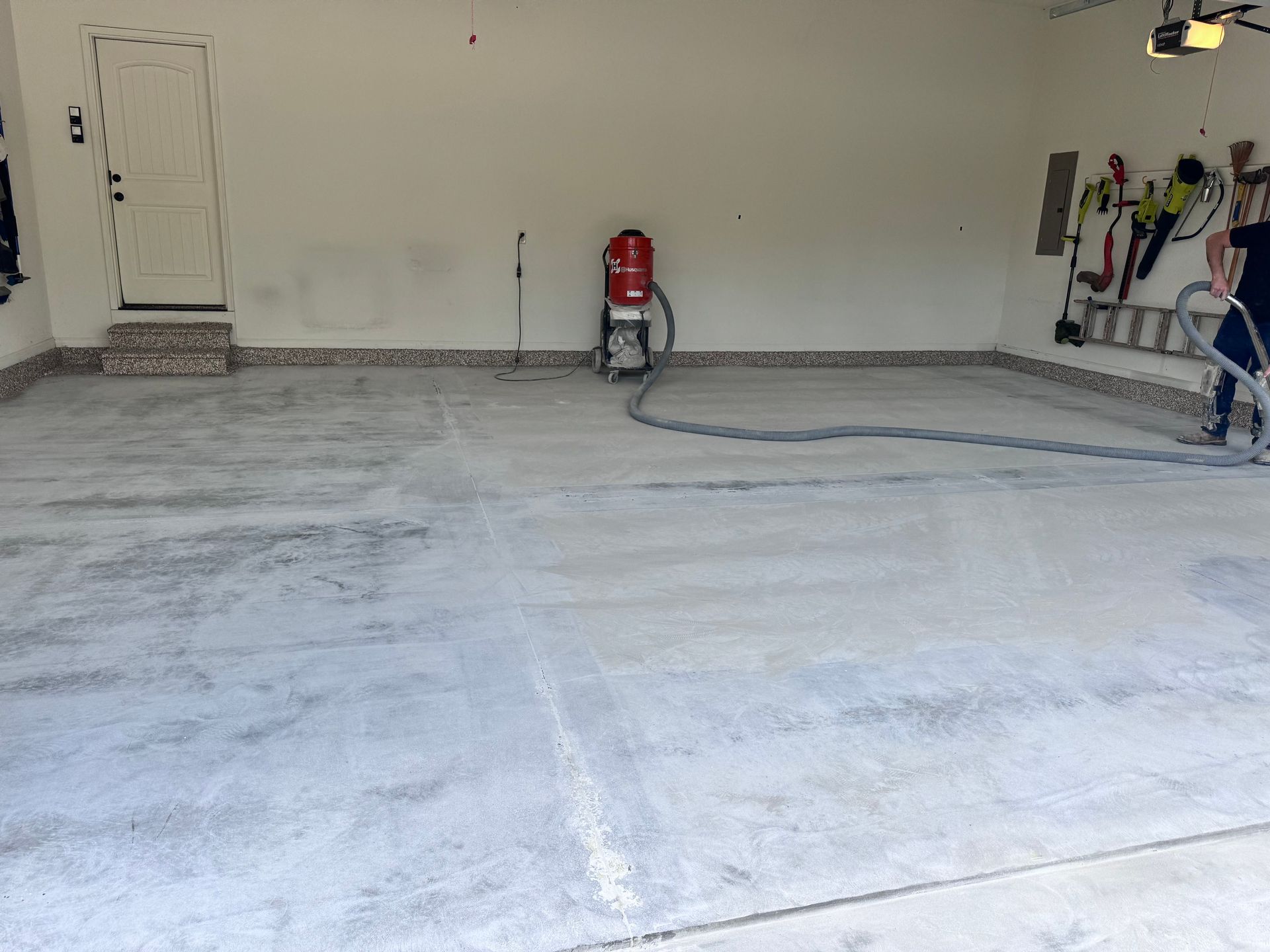 Person using sandblaster on a garage floor; red machine, light gray concrete, white door.