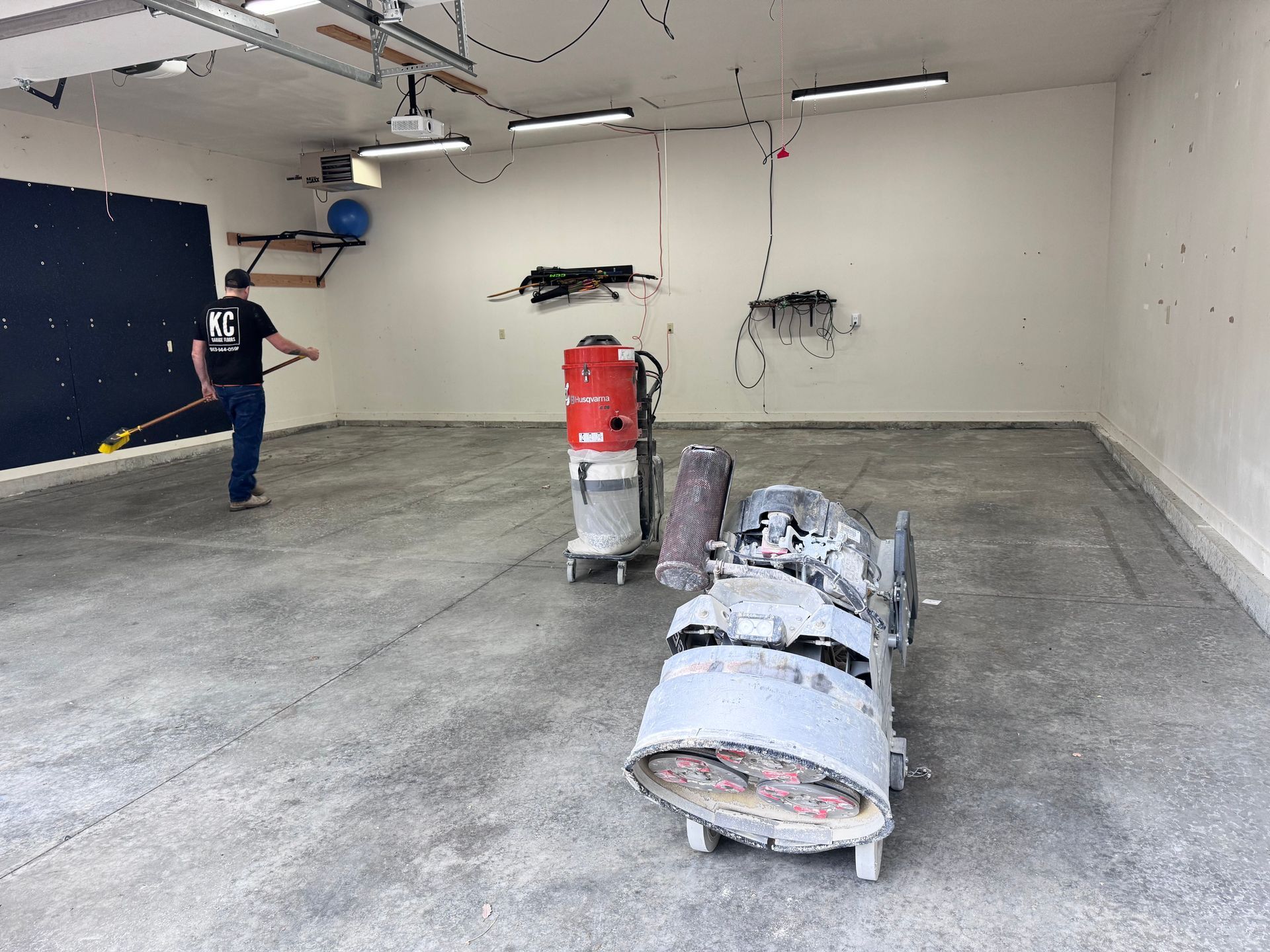 A person sanding a concrete garage floor. Equipment and a vacuum are present.