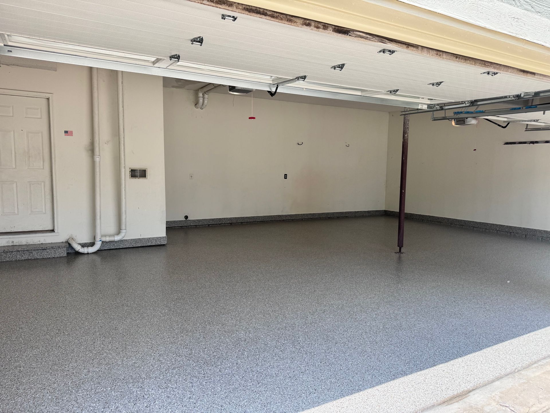 Empty garage with a gray epoxy floor. Beige walls, white door, and overhead garage door opener.