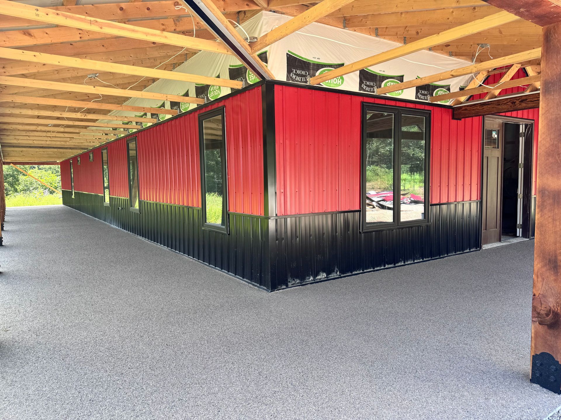 Red and black metal building with windows under a wooden roof, resting on a gray aggregate floor.