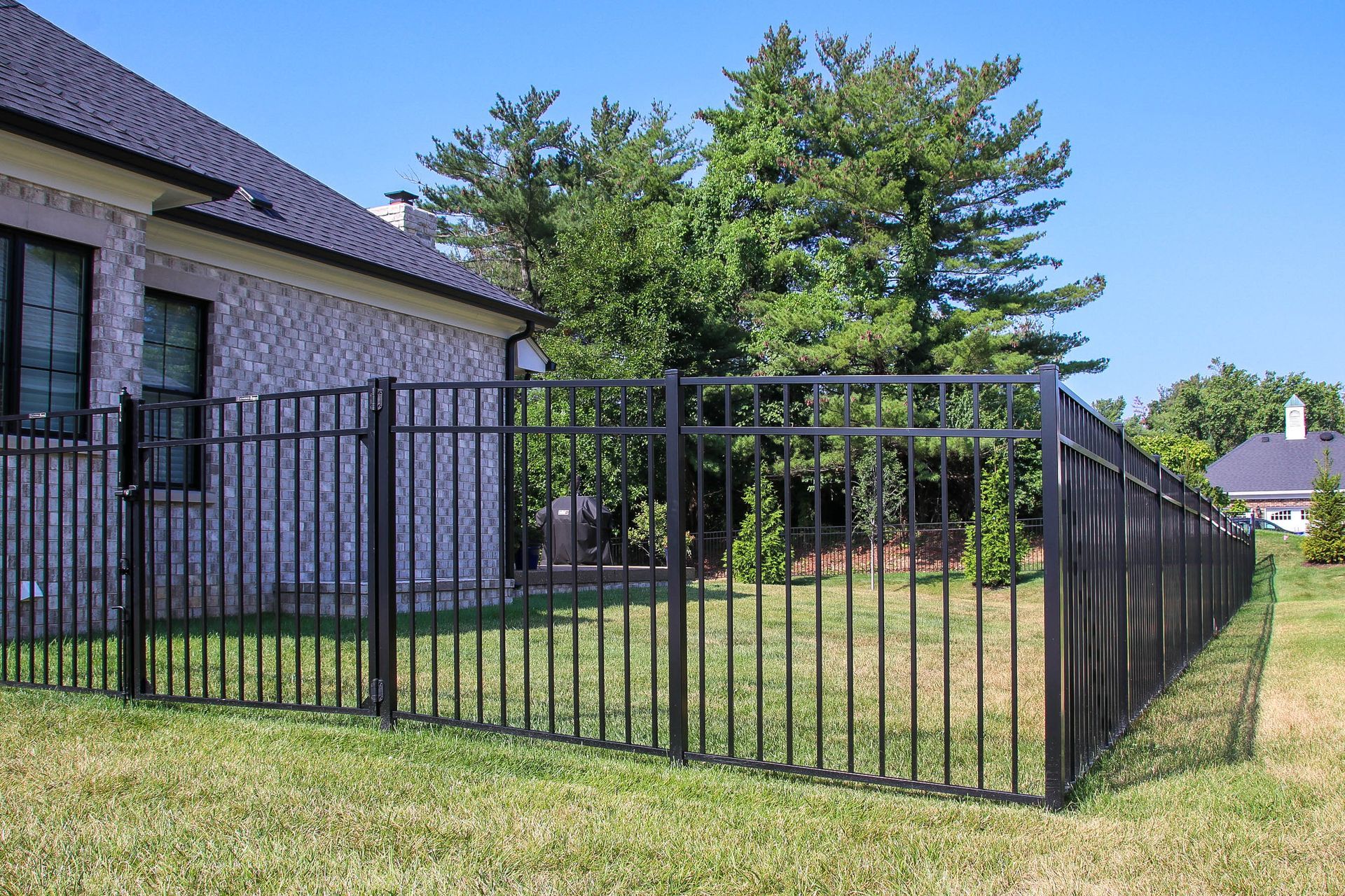 Black metal fence surrounds a grassy yard adjacent to a brick house on a sunny day.