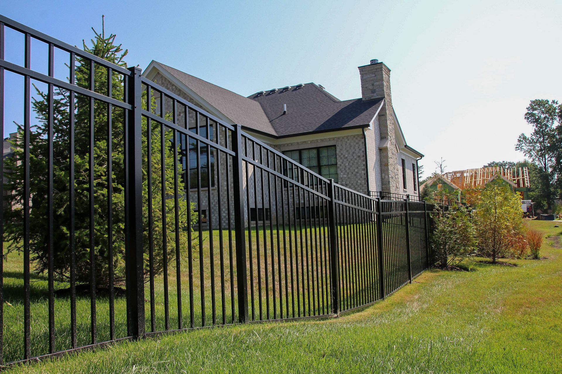 Black metal fence encloses a grassy yard with a house and trees in the background under a clear sky.