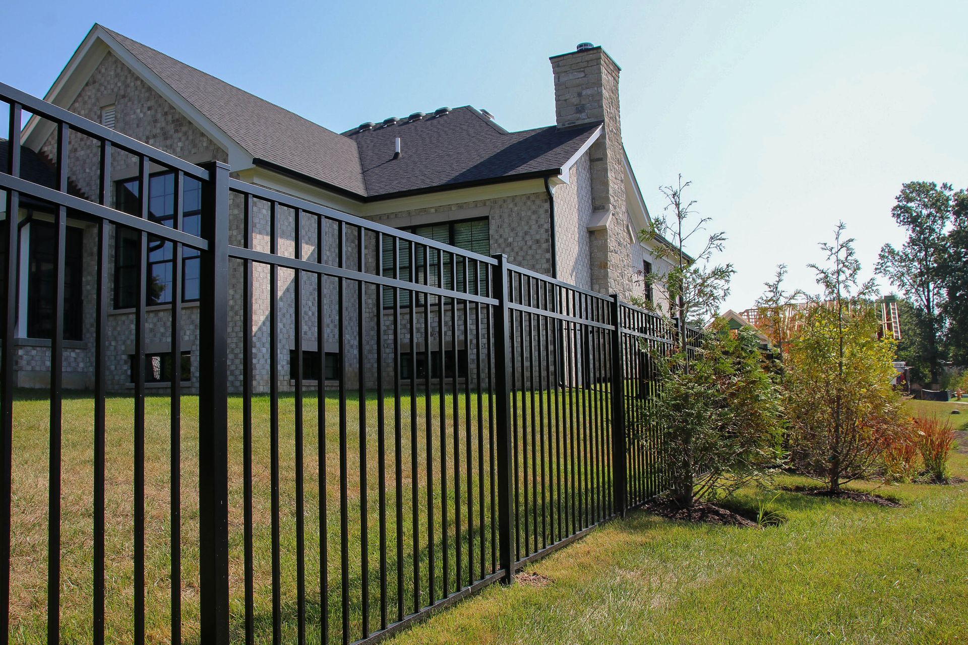 Black metal fence surrounding a brick house with a chimney on a sunny day.