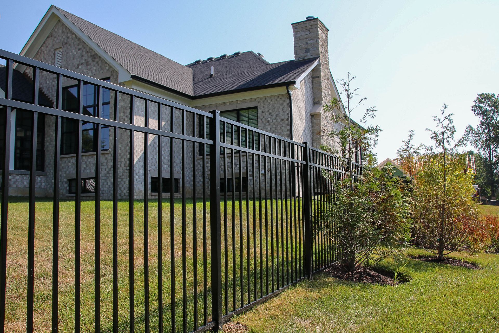 Black metal fence surrounds a light-colored brick house with a chimney on a sunny day.