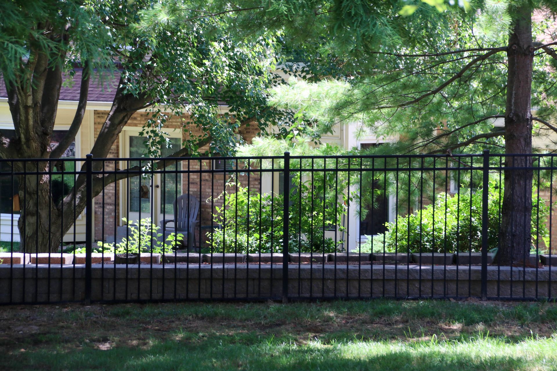 Black metal fence in front of a brick house, partially obscured by trees and greenery.