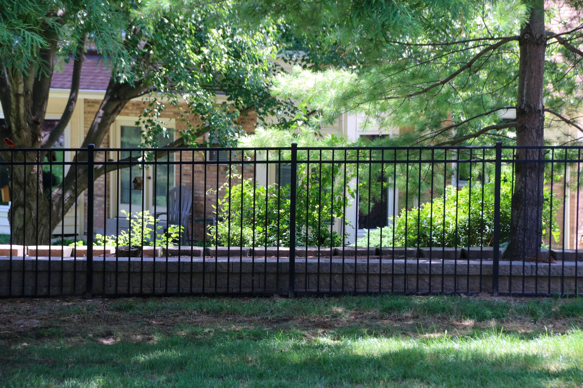 Black metal fence in front of a brick wall and green foliage, with a house visible in the background.