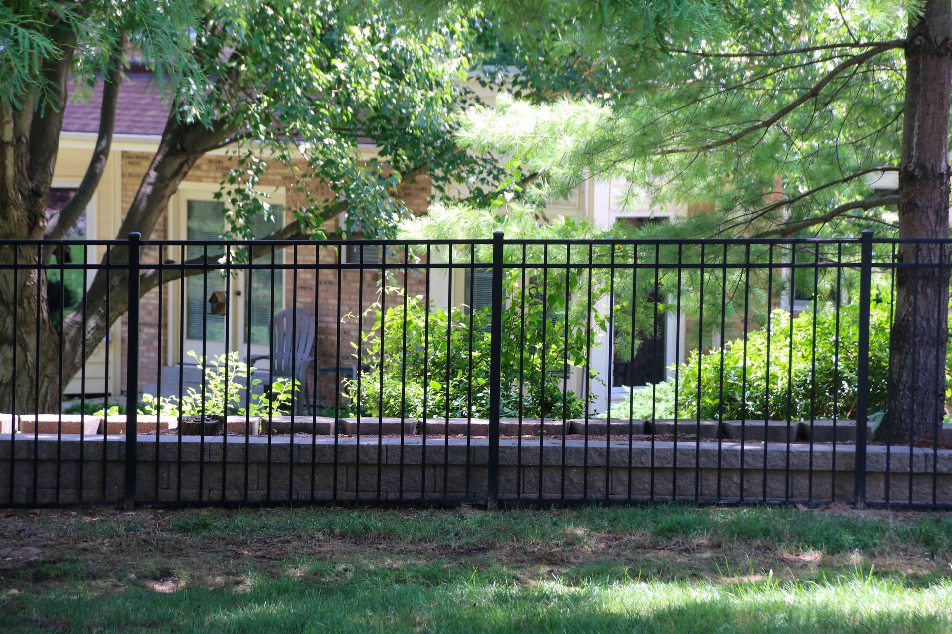 Black metal fence atop a low stone wall, with a house and trees in the background.