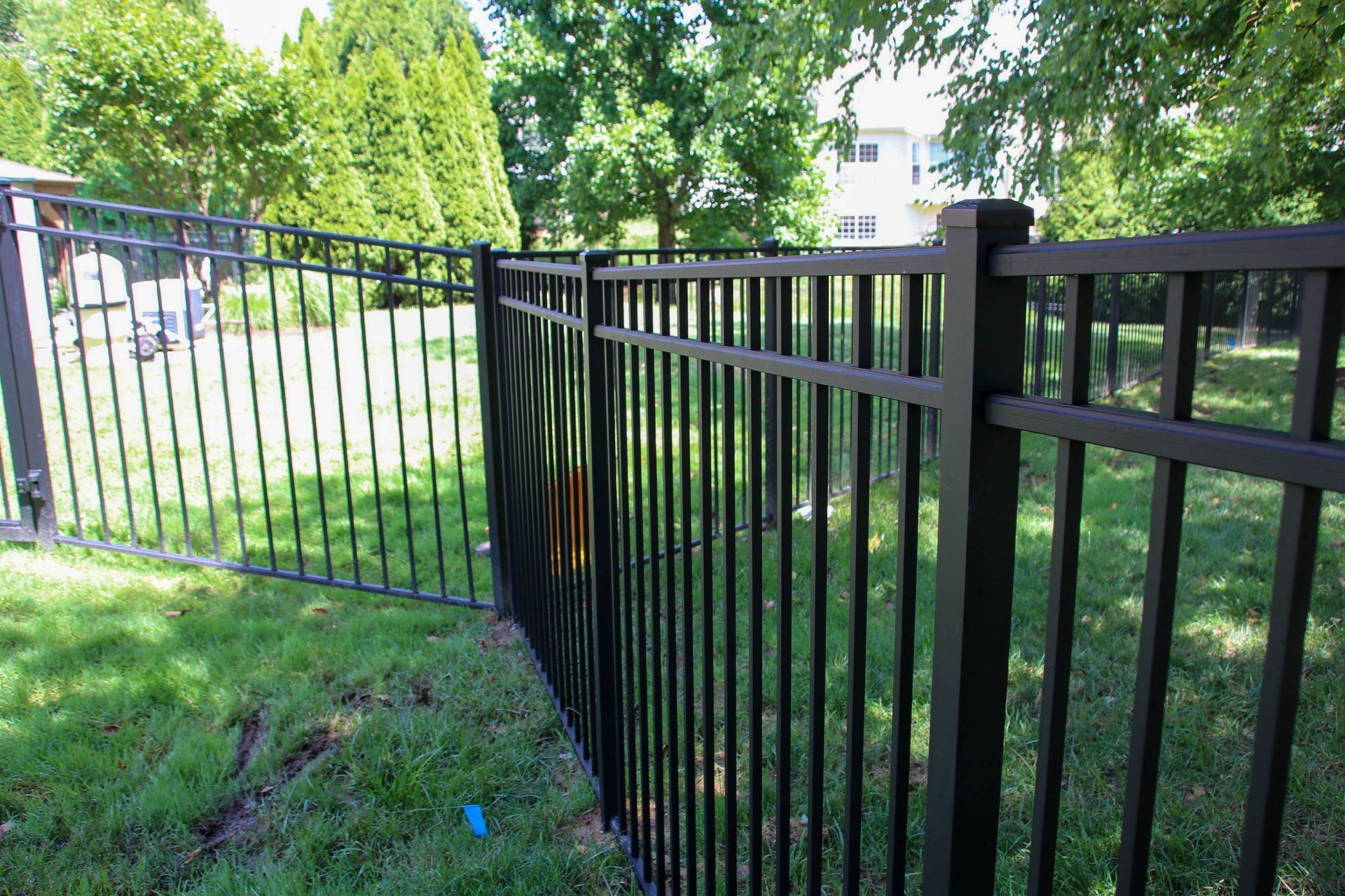 Black metal fence in a grassy yard, with a gate, trees and sky in the background.