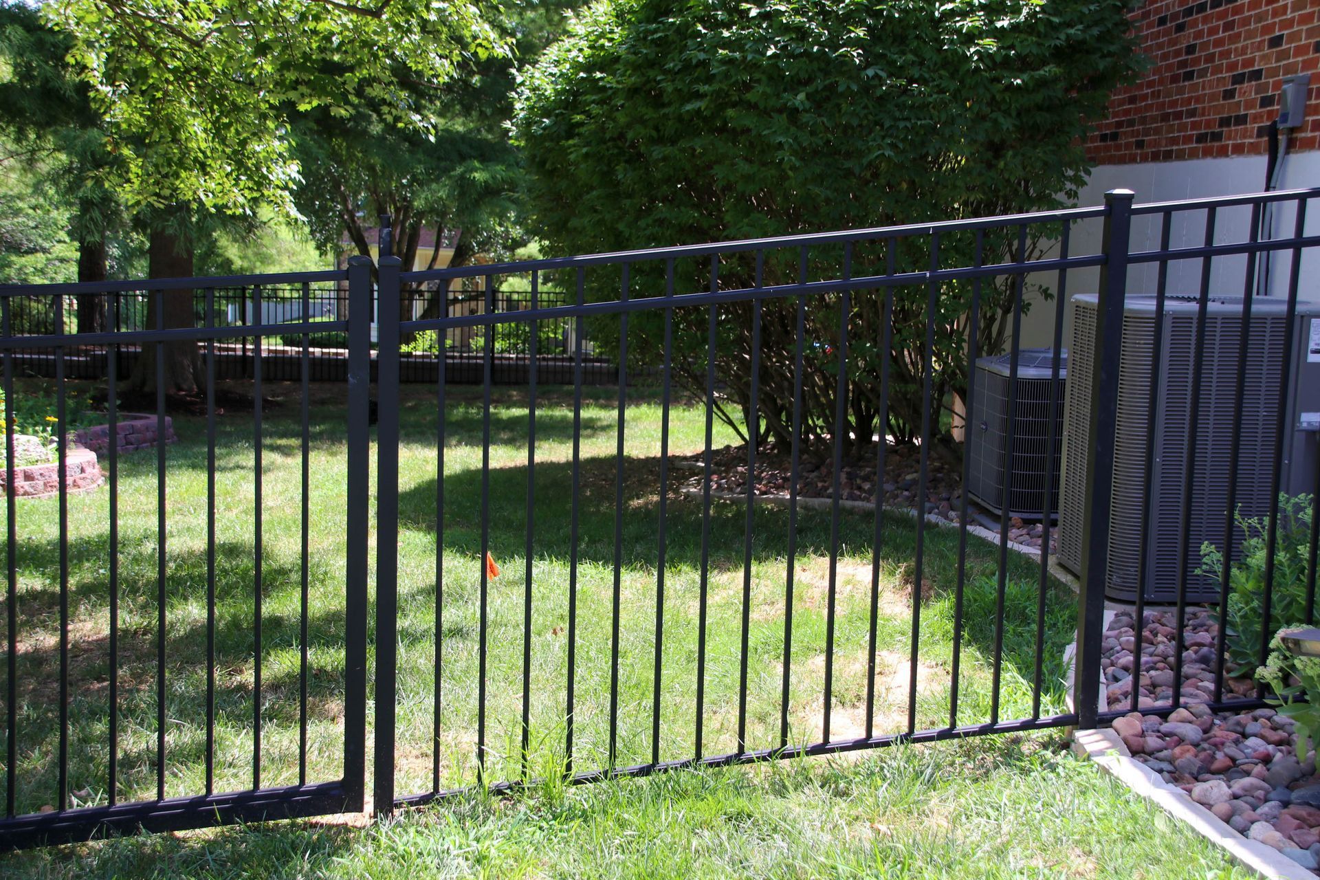 Black metal fence encloses a grassy backyard with a tree, bushes, and an air conditioning unit.