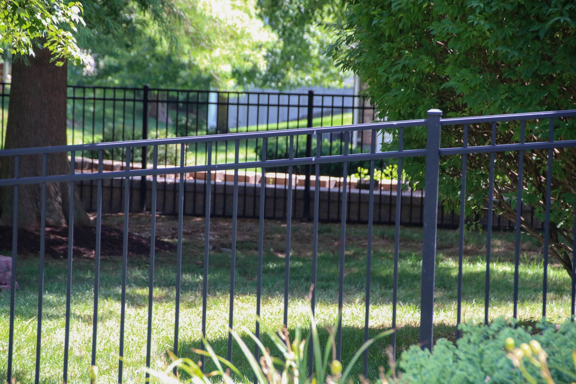 Black metal fence in a yard with trees and a brick retaining wall.