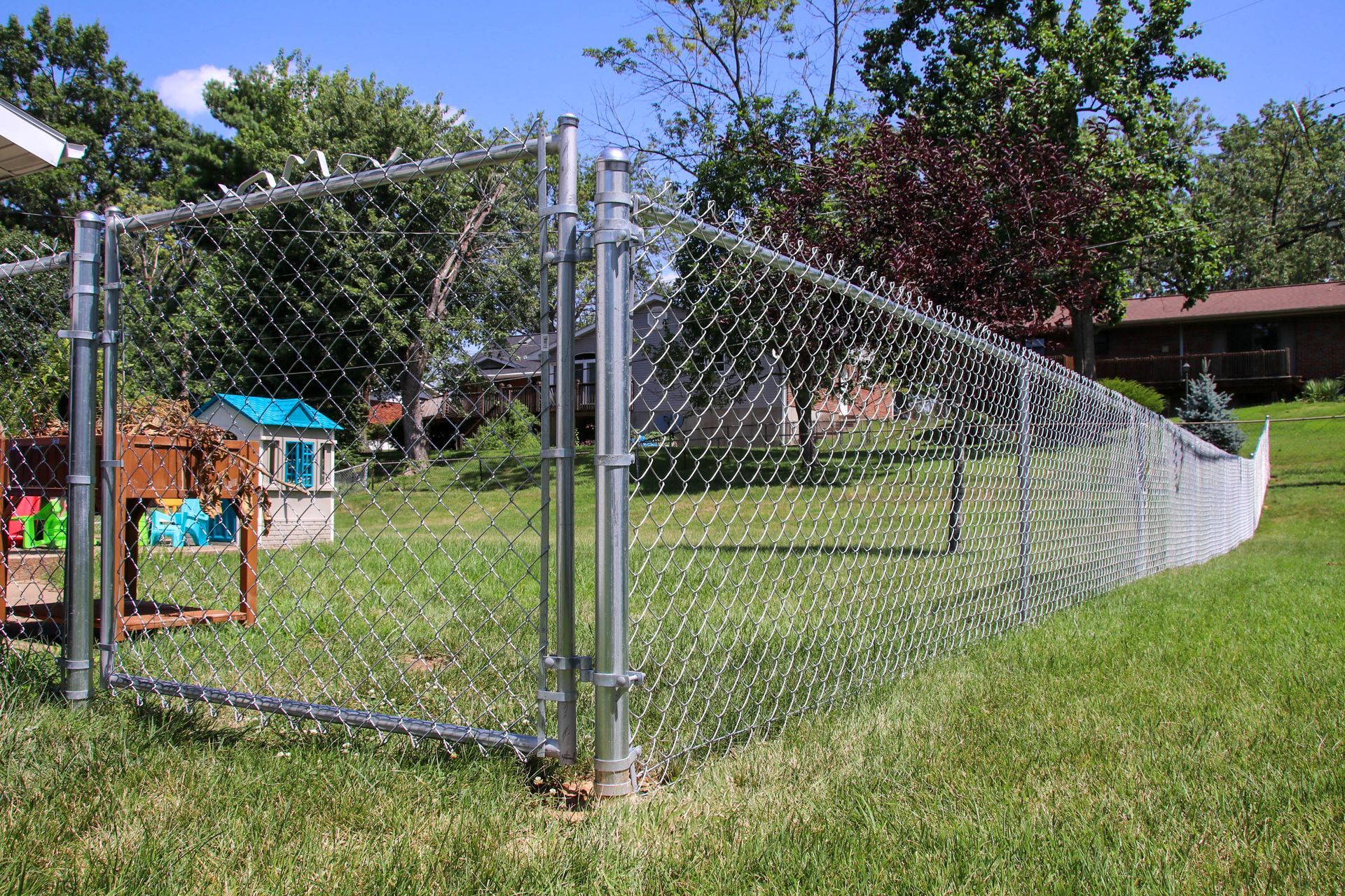 Chain-link fence encloses a grassy backyard with a playhouse and trees under a blue sky.
