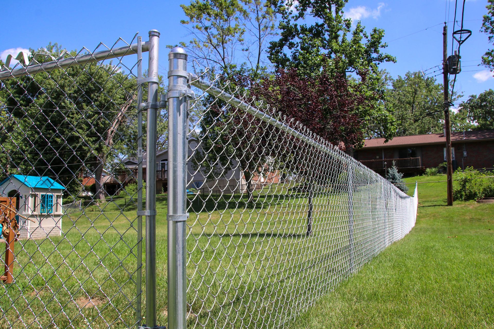 Chain-link fence in a backyard, surrounding a grassy area with a playhouse and trees in the background.