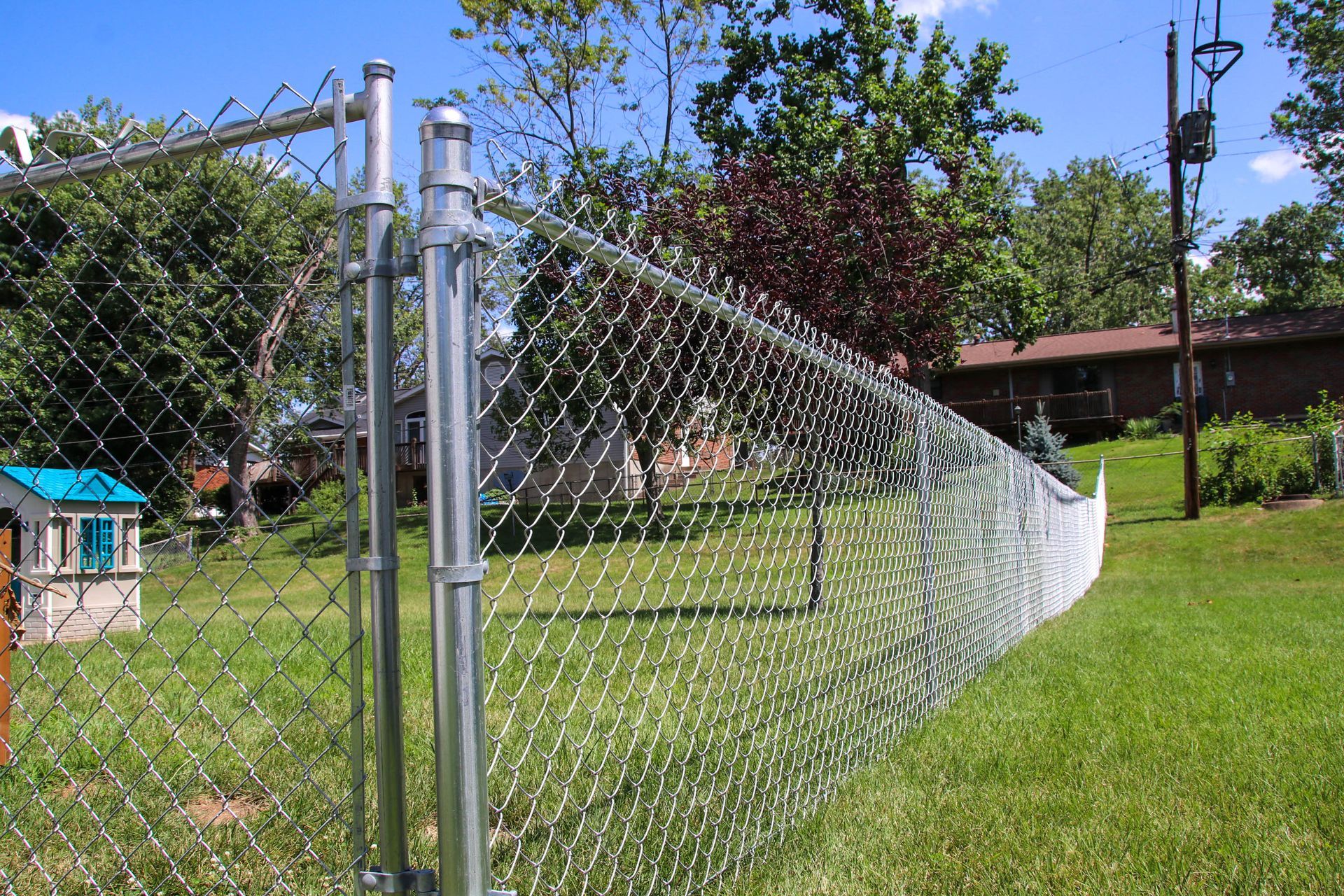 Chain-link fence in a grassy yard, with a small playhouse and houses in the background on a sunny day.
