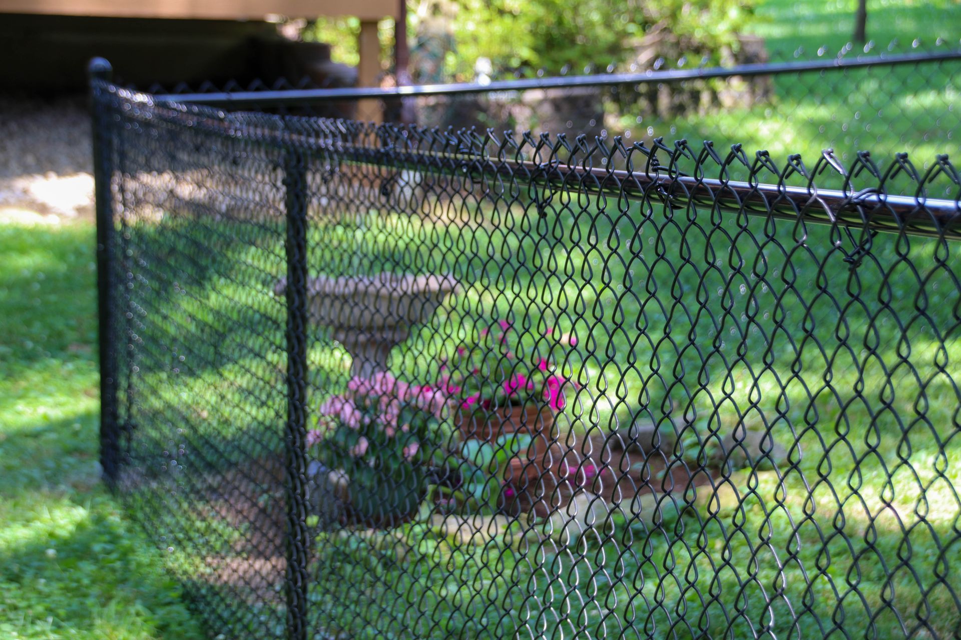 Black chain link fence encloses a garden with a birdbath and pink flowers, in a grassy yard.