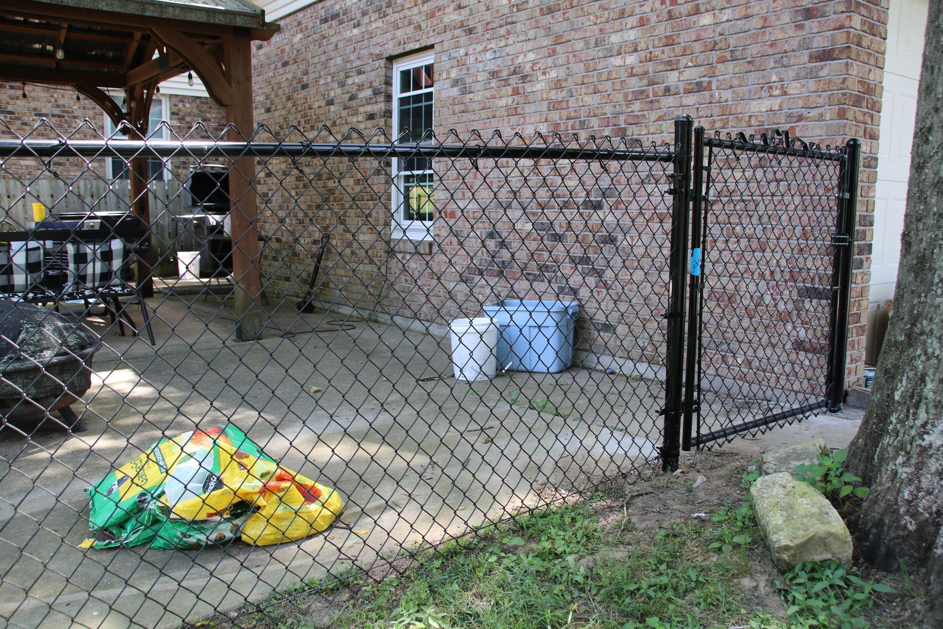 Black chain-link fence with gate, against a brick building. Two buckets and a colorful toy bag sit inside.