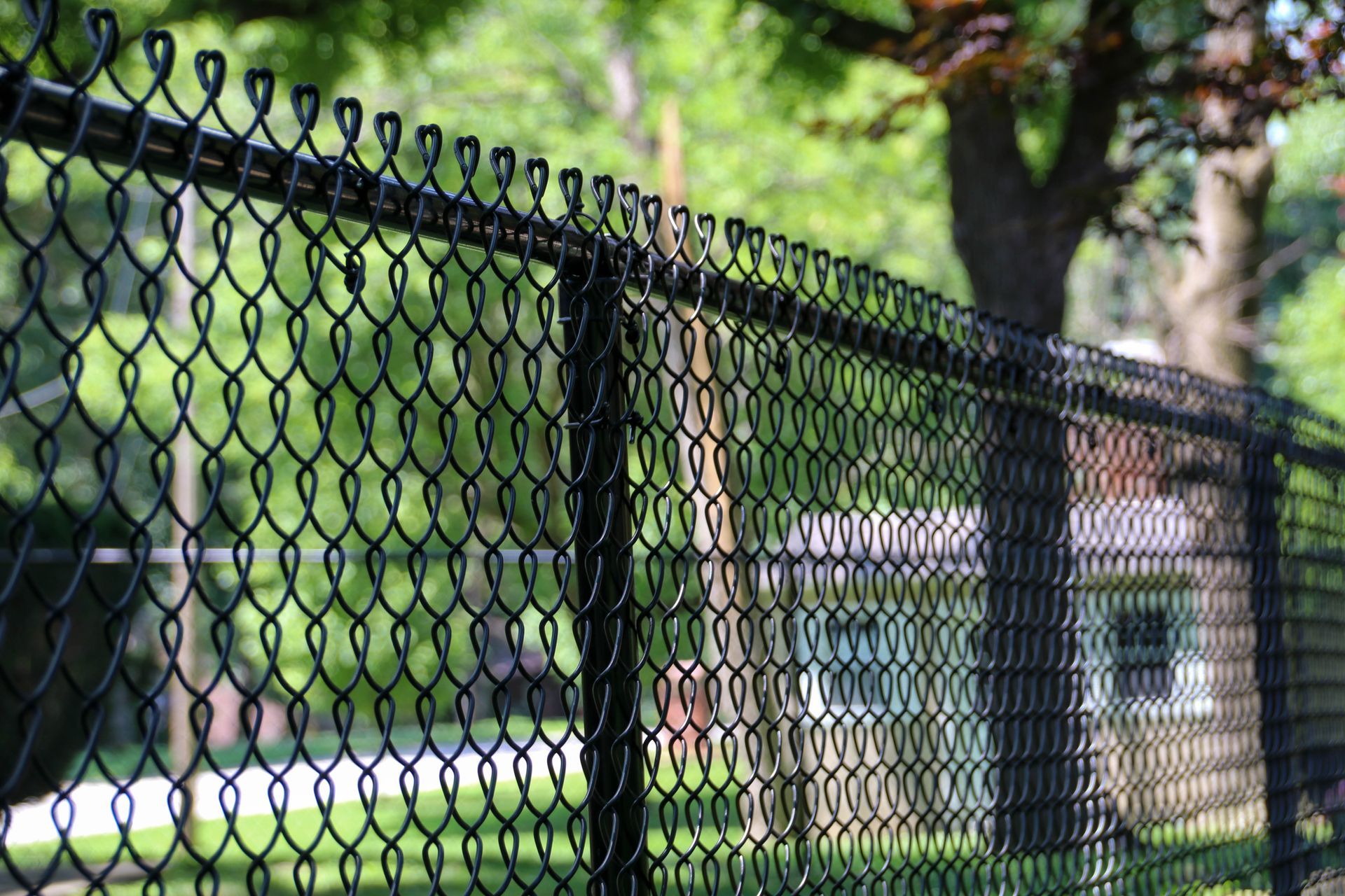 Black chain-link fence with a house and trees blurred in the background on a sunny day.
