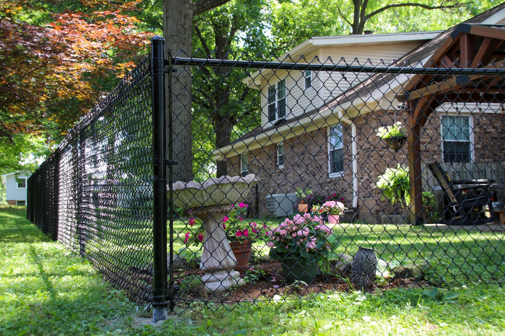 Black chain-link fence surrounds a backyard garden with a birdbath and flowering plants near a brick house.