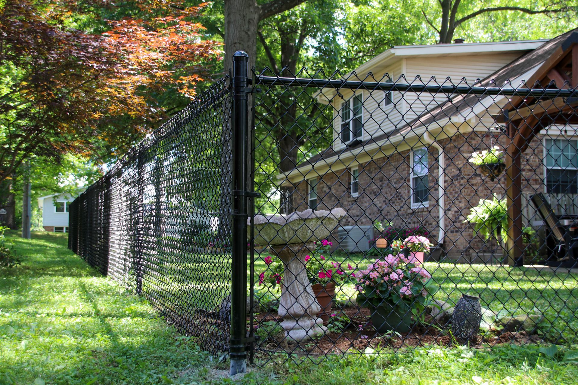 Black chain-link fence in front of a house with a garden and bird bath. Lush green grass and trees in background.