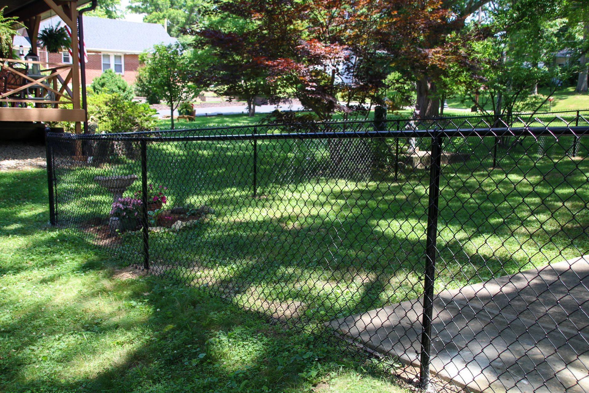 Black chain link fence in a grassy yard, partially enclosing a flower bed. A deck and trees are in the background.