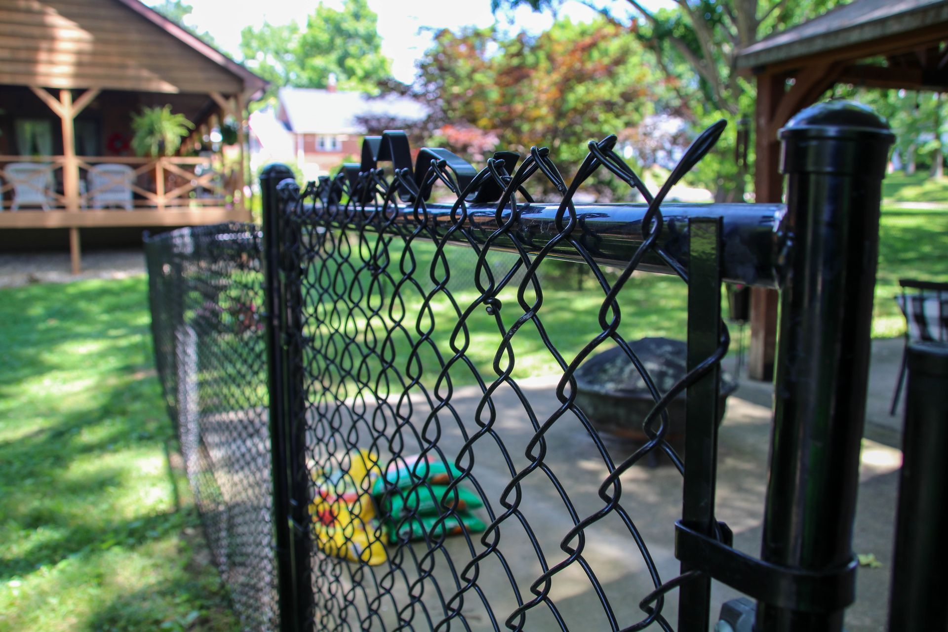 Black chain-link fence in focus; background features houses and gazebo. Green lawn, sunny day.