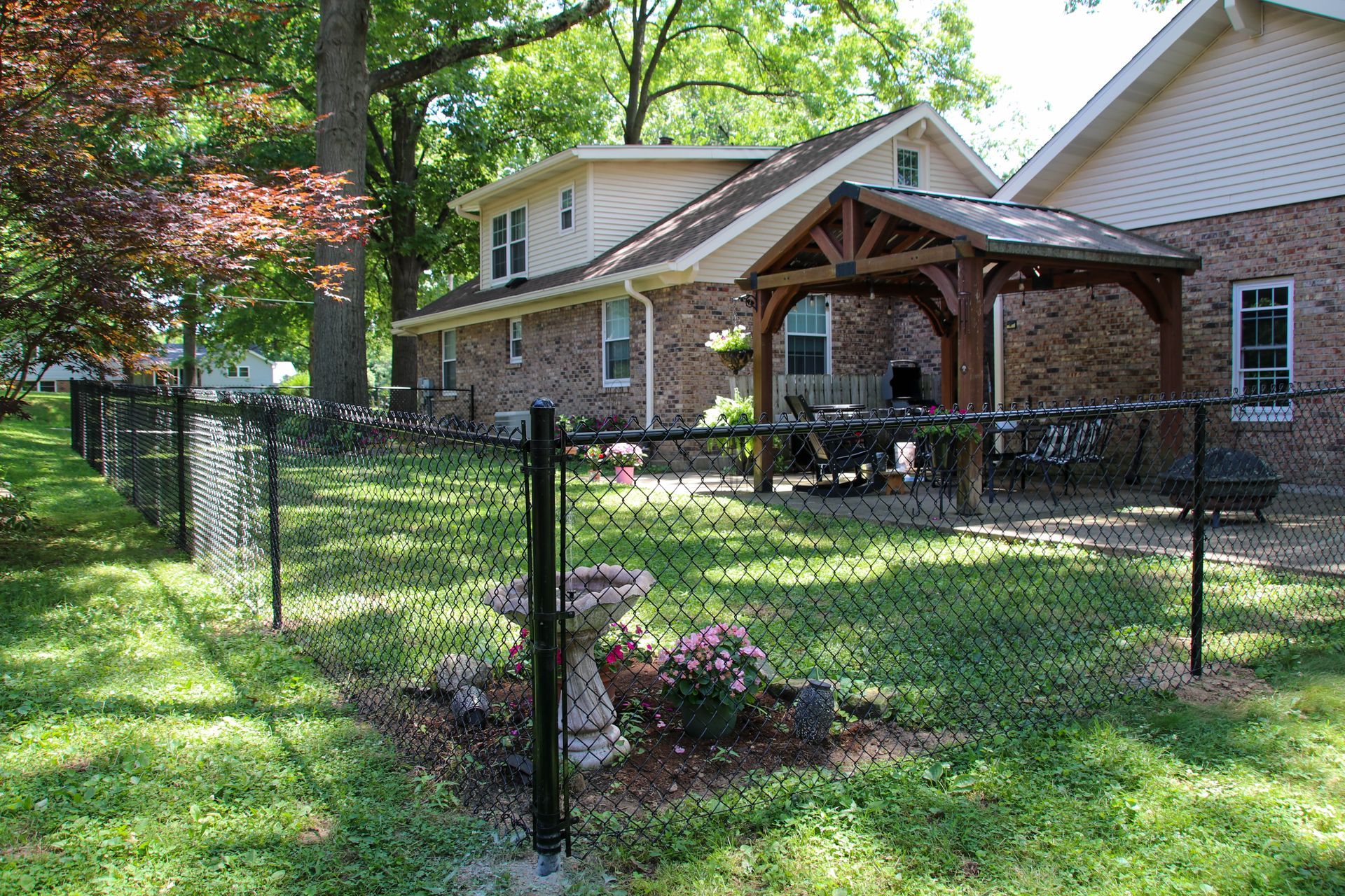 Black chain-link fence surrounds a backyard with a brick house and gazebo. Green grass and trees are in the setting.