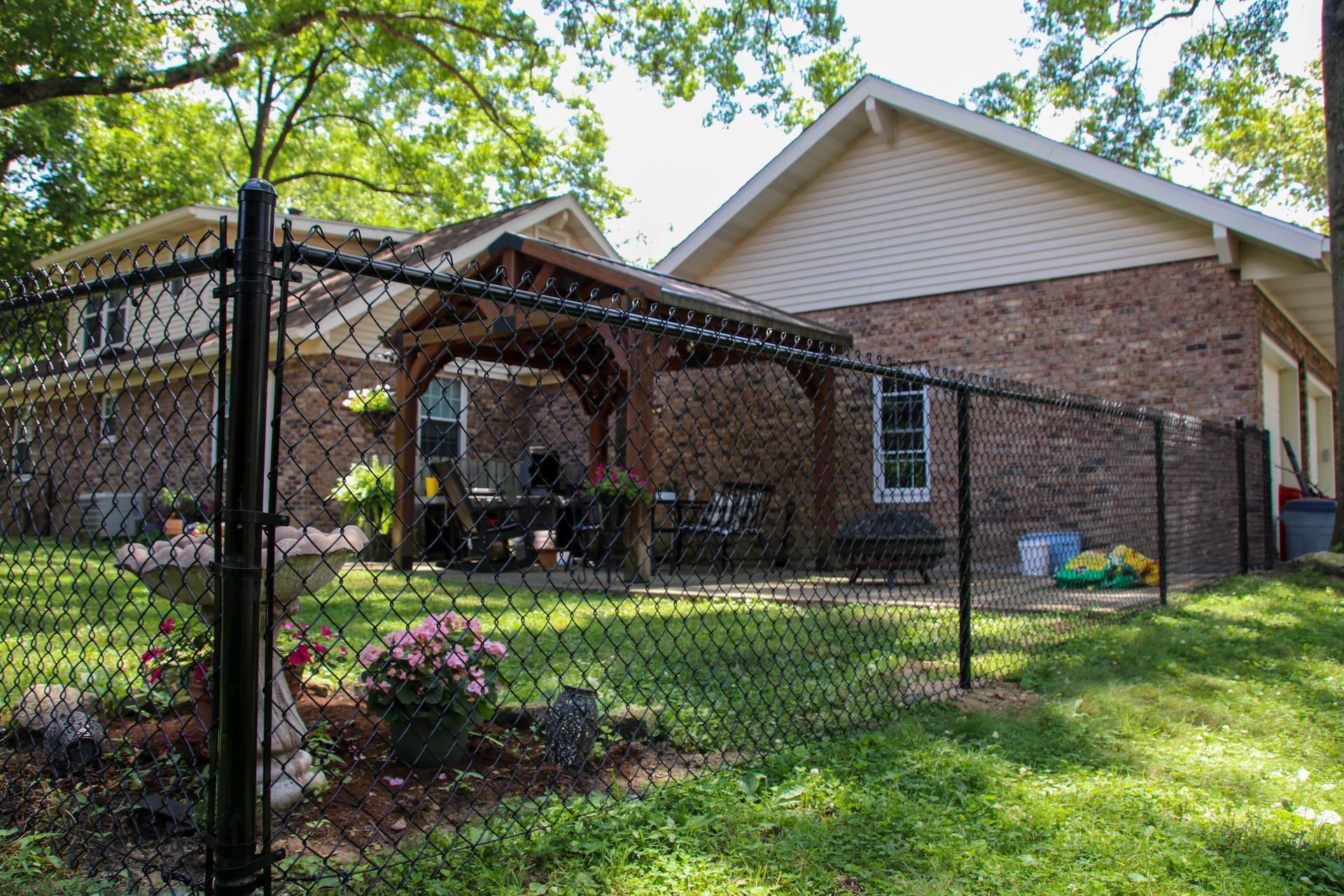 Black chain-link fence surrounds a brick house with a covered patio, flowers, and greenery.