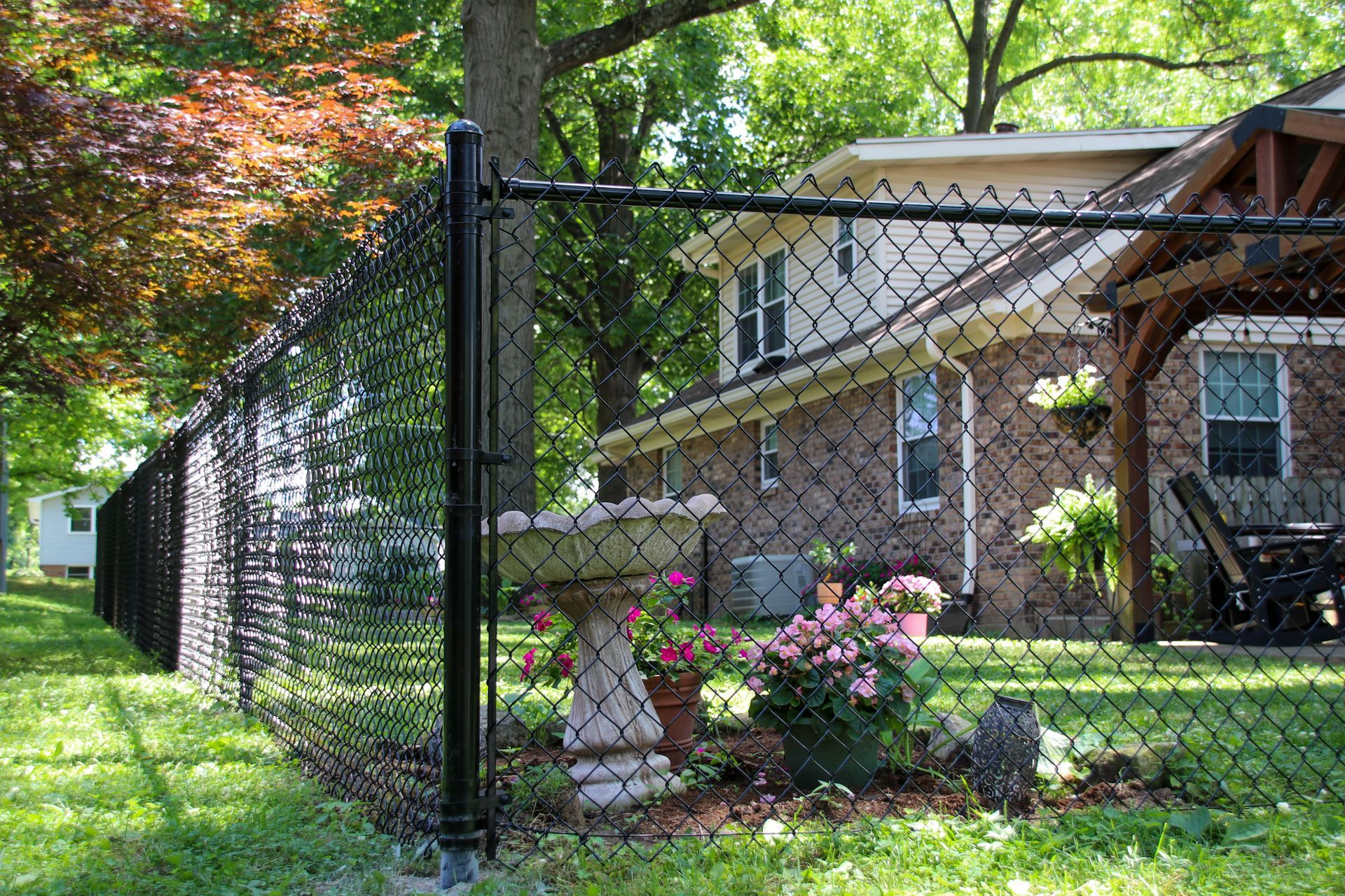 Black chain-link fence in front of a house with a gazebo, bird bath, and flowers.