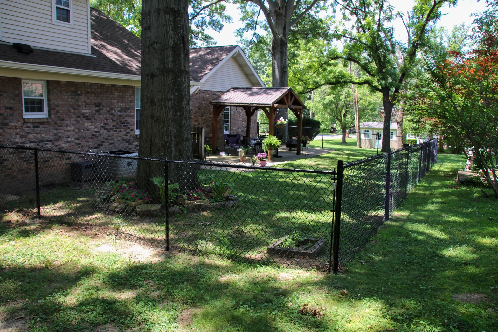 Black chain-link fence in backyard, brick house left, porch in background. Trees and green grass surround.