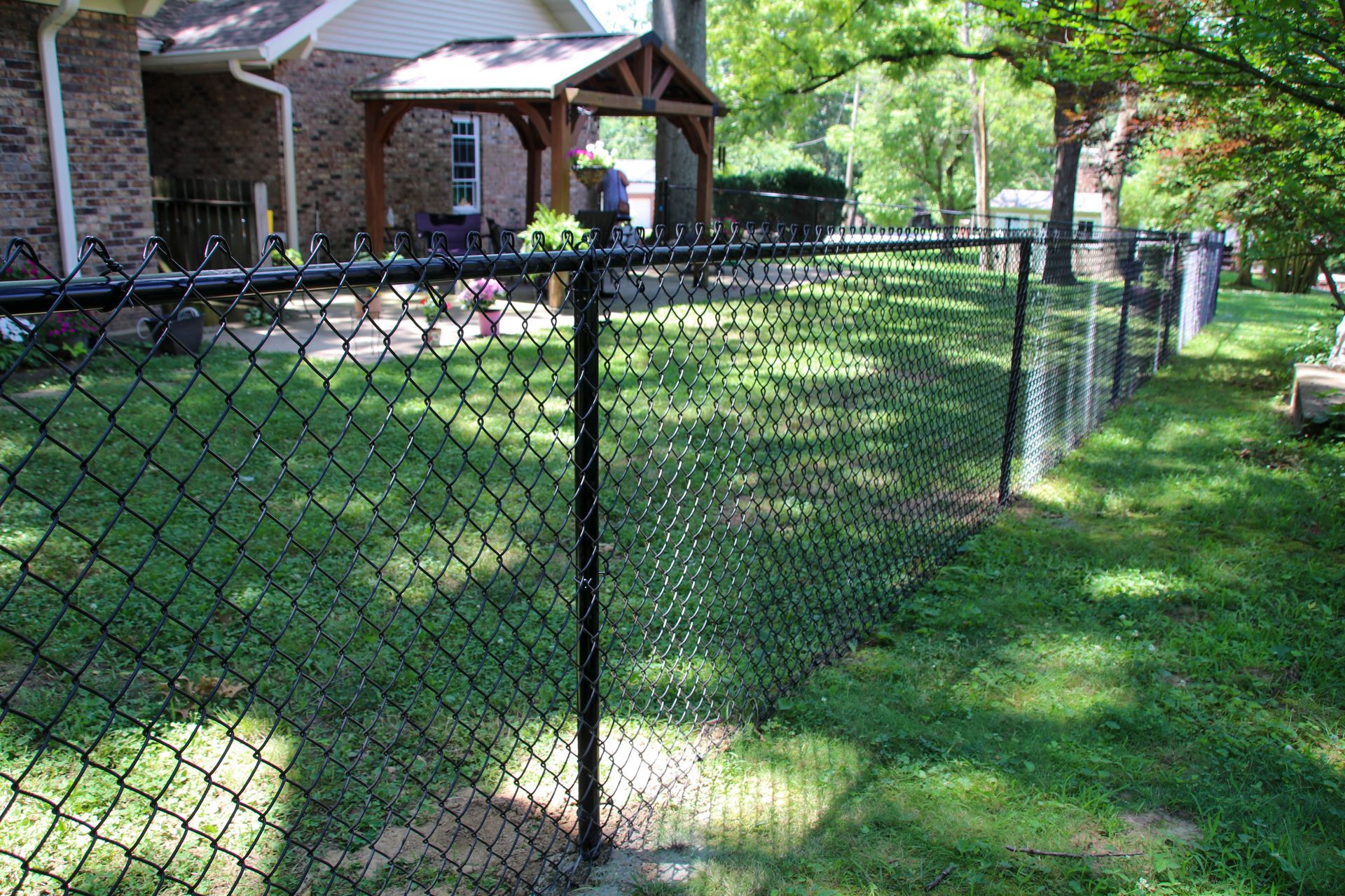 Black chain-link fence in a grassy yard, with a brick house and wooden porch in the background.