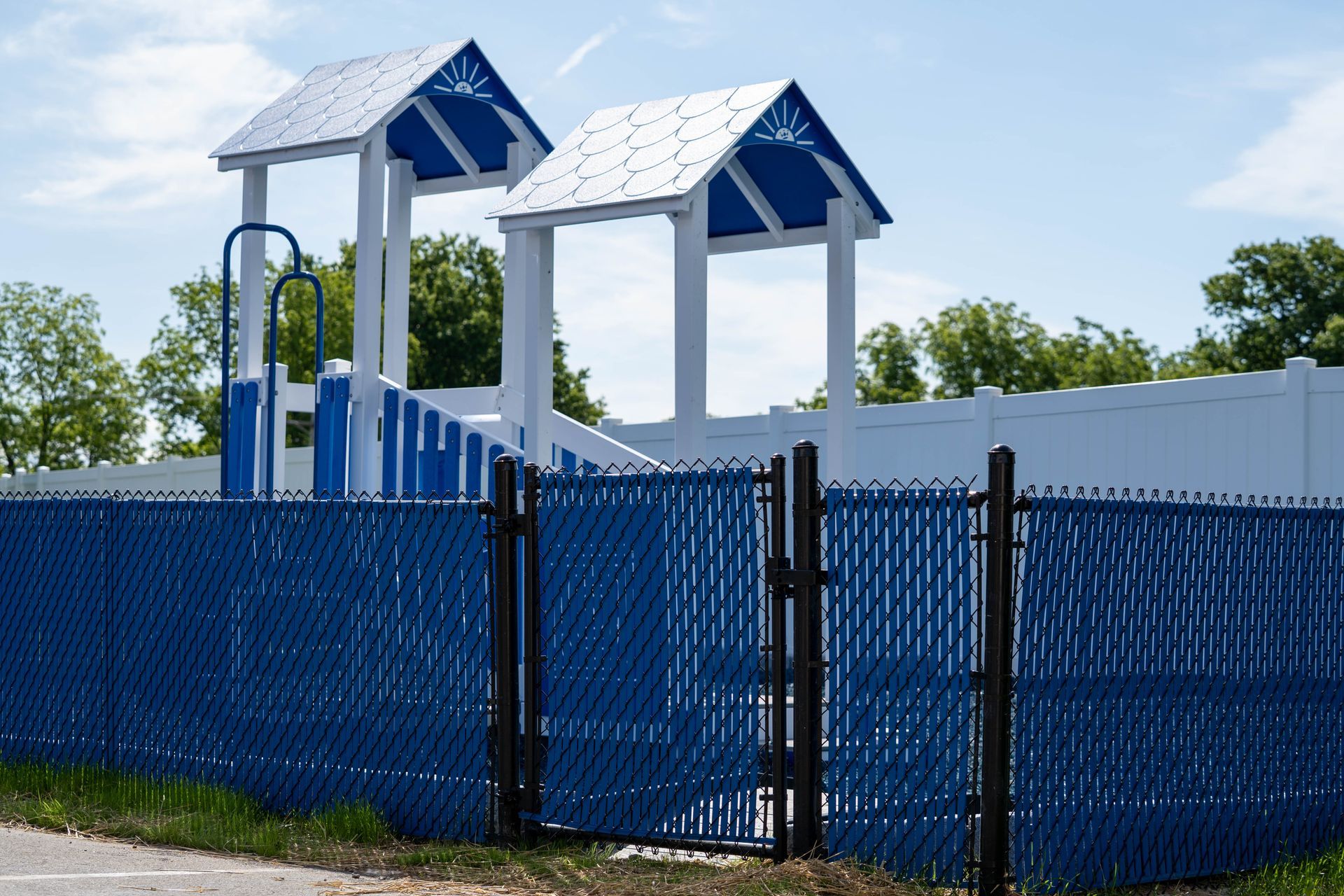 Blue fenced playground with two blue-roofed structures, white frames, and a blue gate.