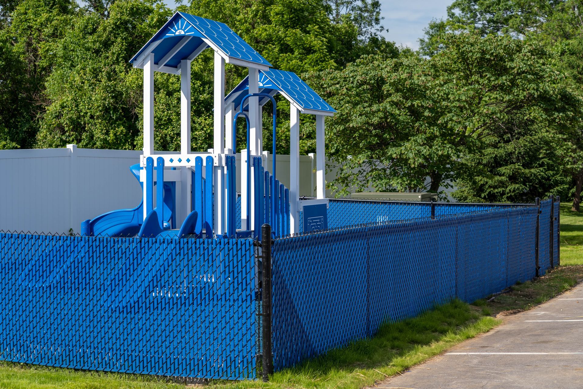 Blue playground equipment inside a blue privacy fence, set in a park with trees.