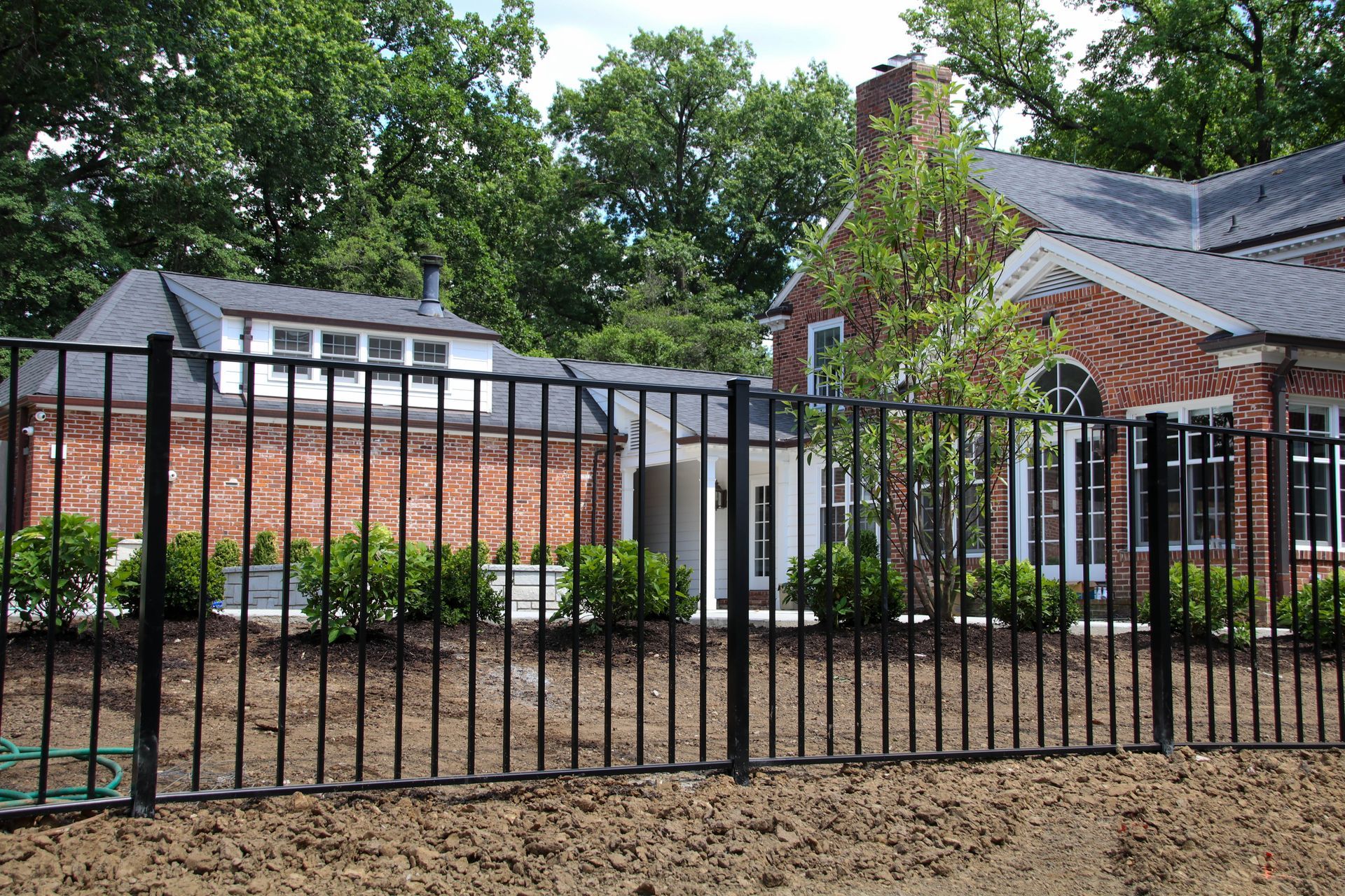 Black metal fence in front of a brick house with a dark roof and lush green trees in the background.