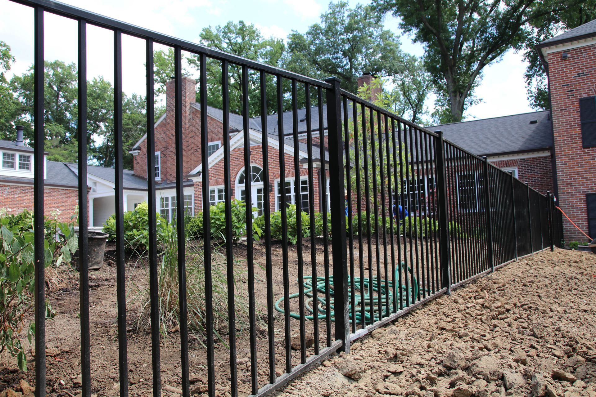Black metal fence in front of a brick building. Dirt and greenery are visible behind the fence.