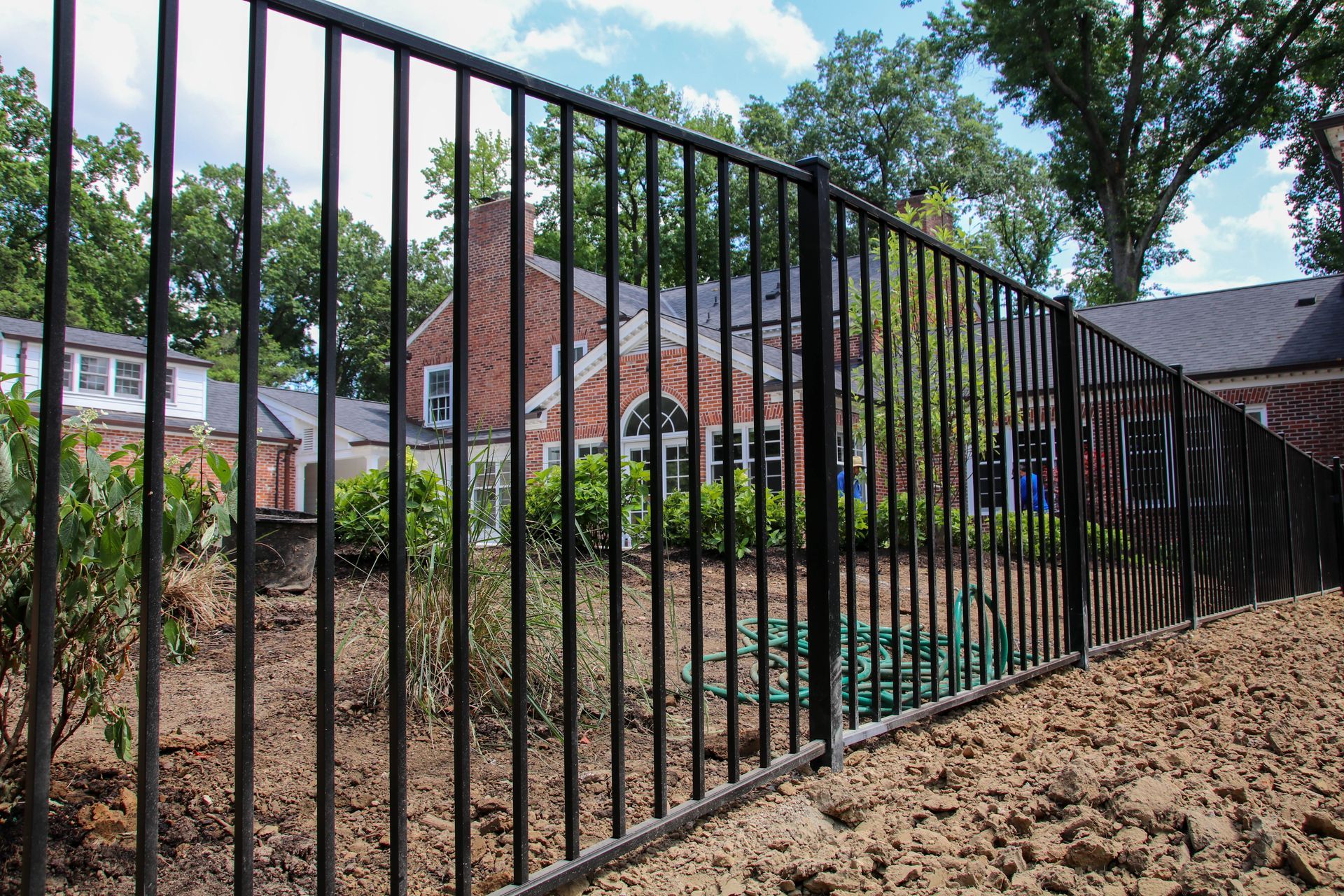 Black metal fence in front of a brick building and trees on a sunny day.