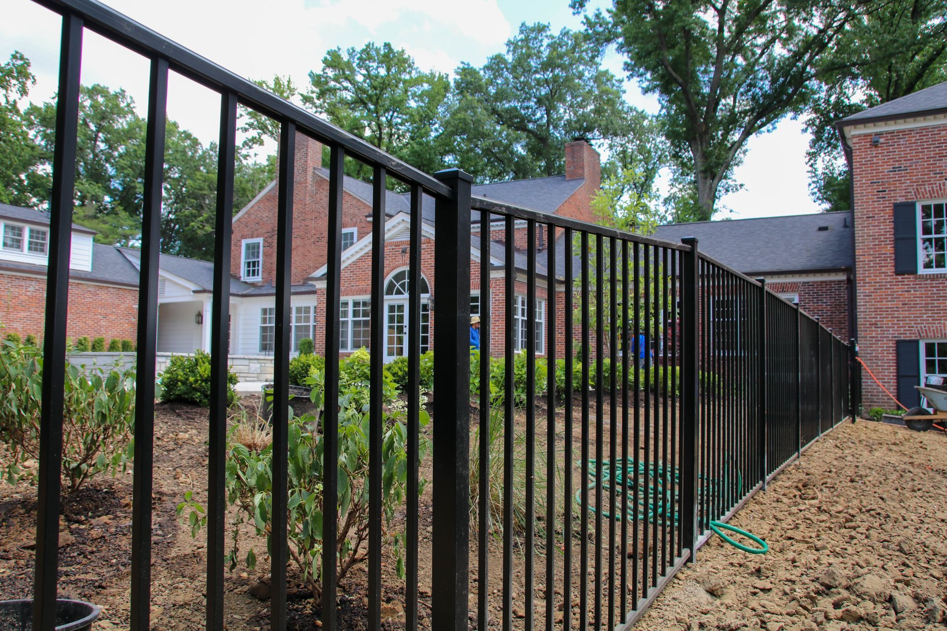 Black metal fence in front of a brick building and landscaping, under a partly cloudy sky.