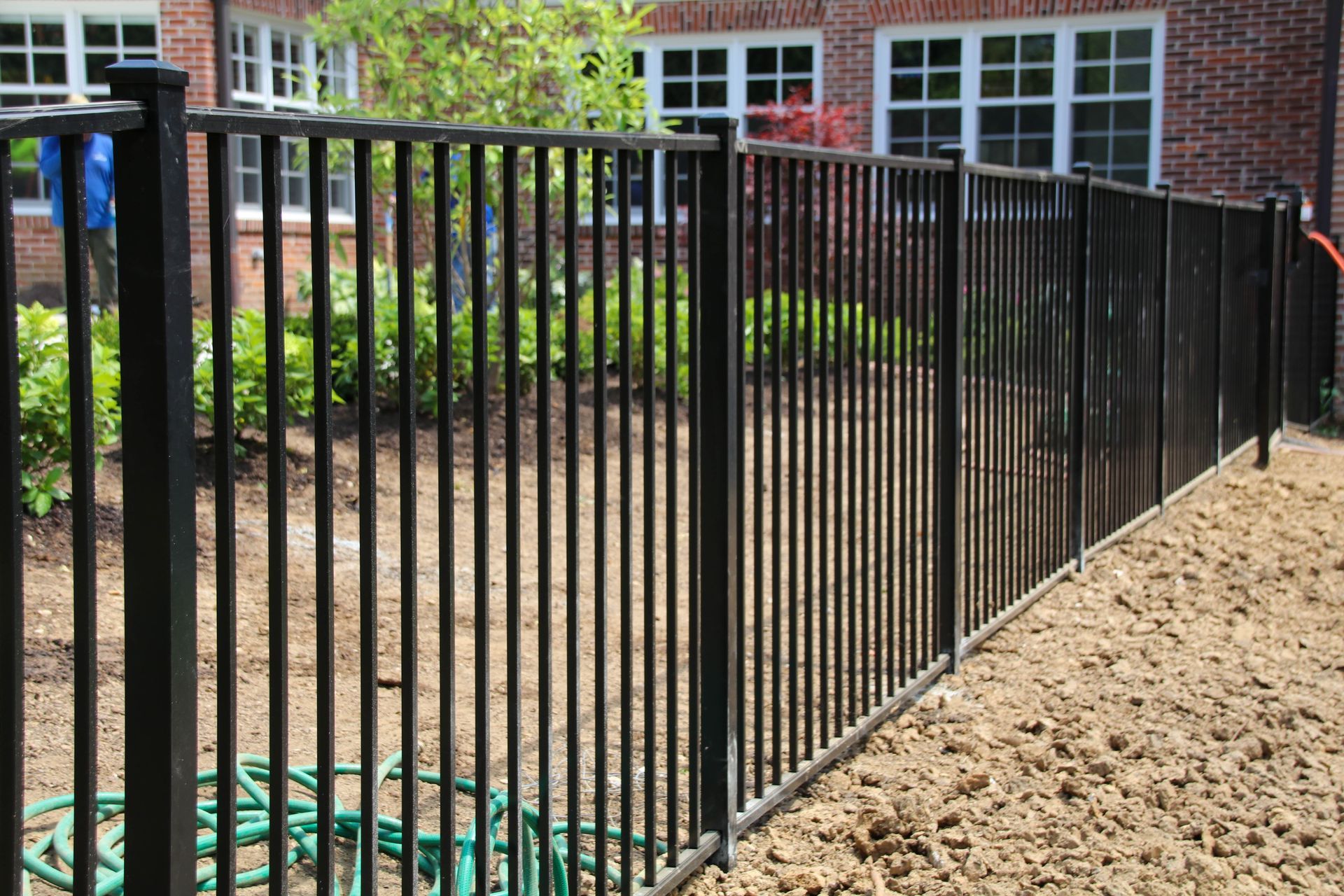Black metal fence surrounding a yard with red brick building and green plants.