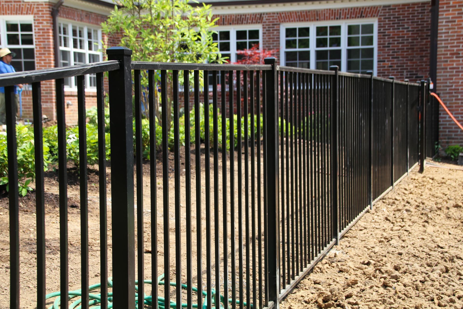 Black metal fence in front of a brick building and garden with green plants.