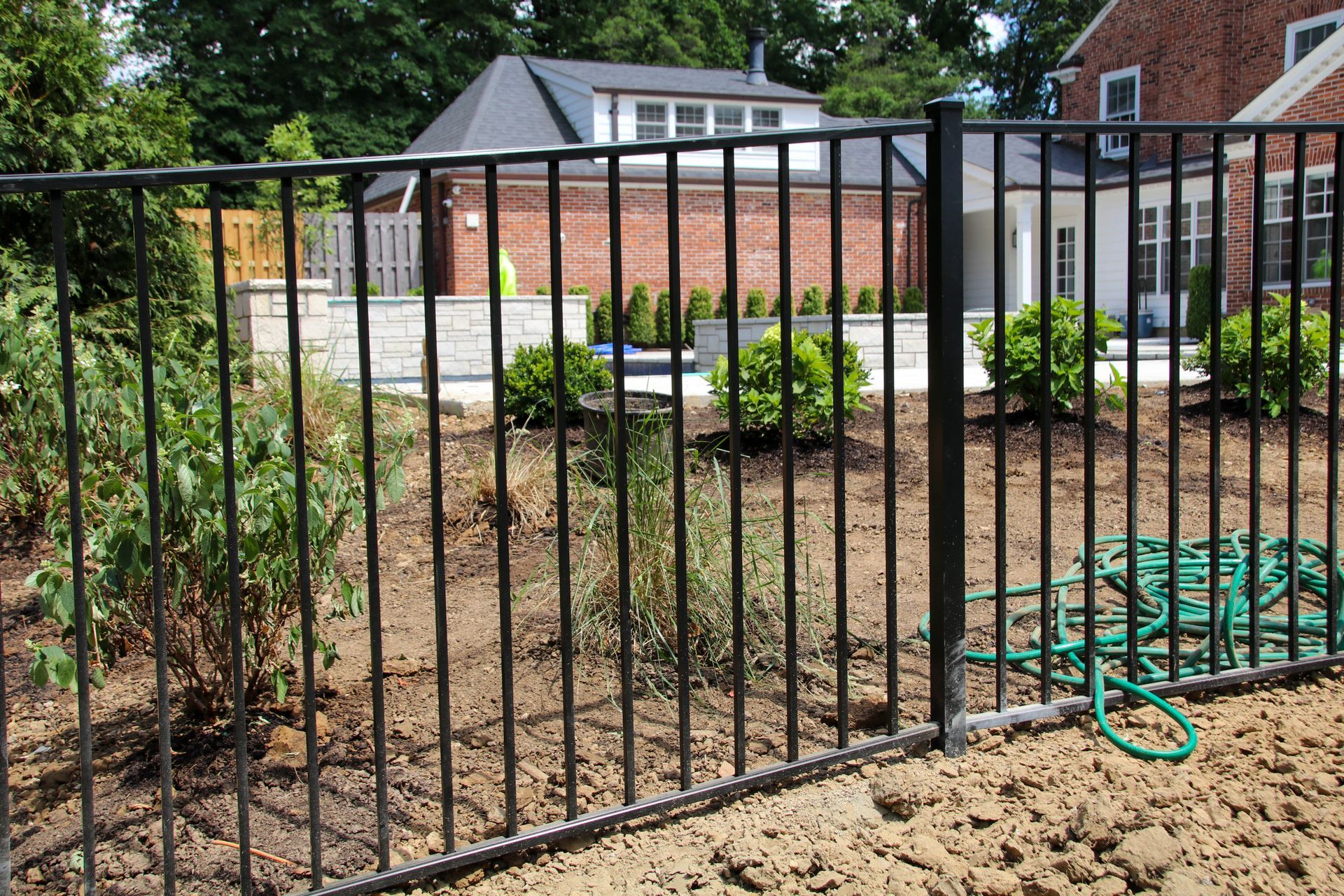 Black metal fence surrounding a garden with bushes, and a brick house in the background.