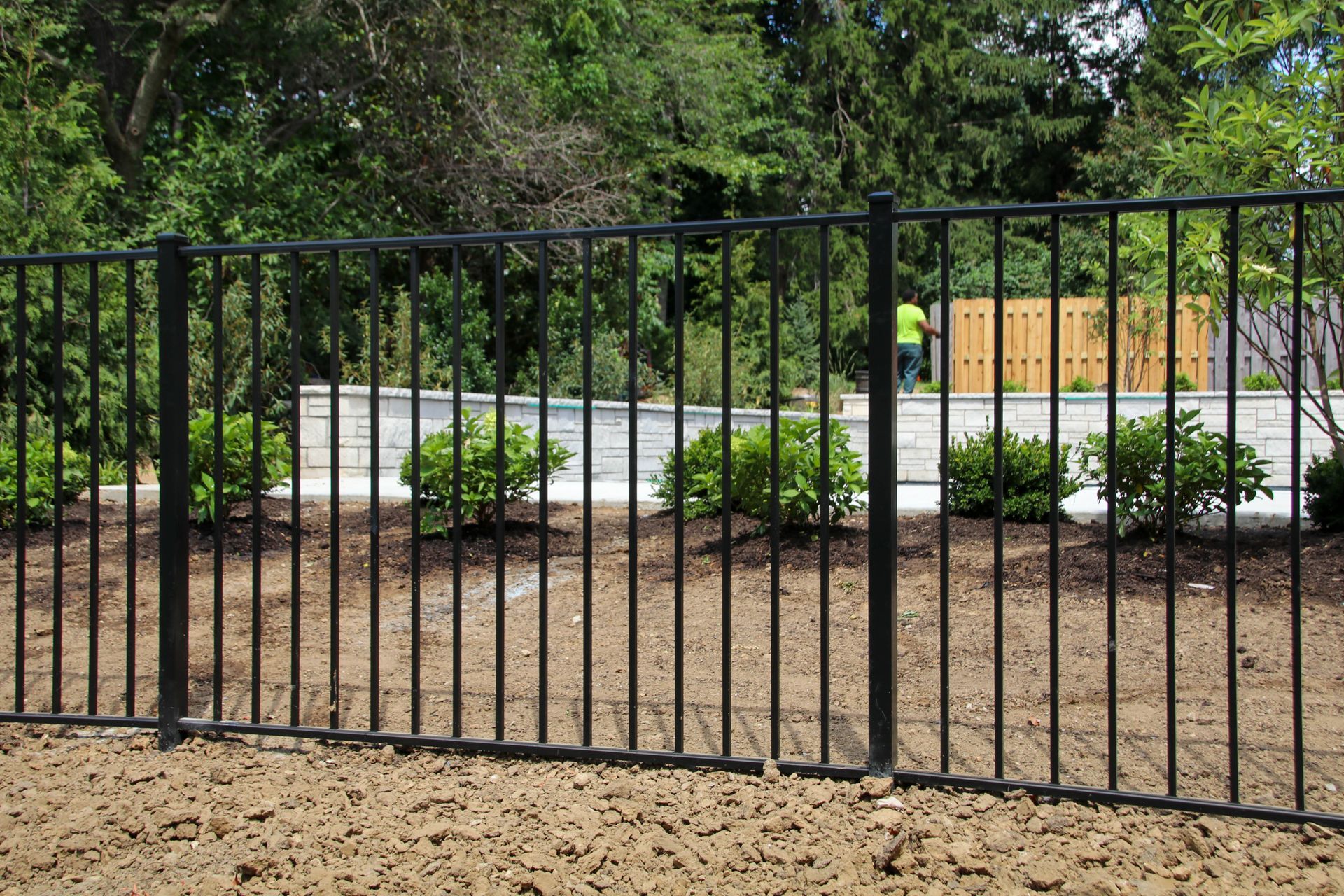 Black metal fence in front of green bushes, with a white wall and trees in the background.