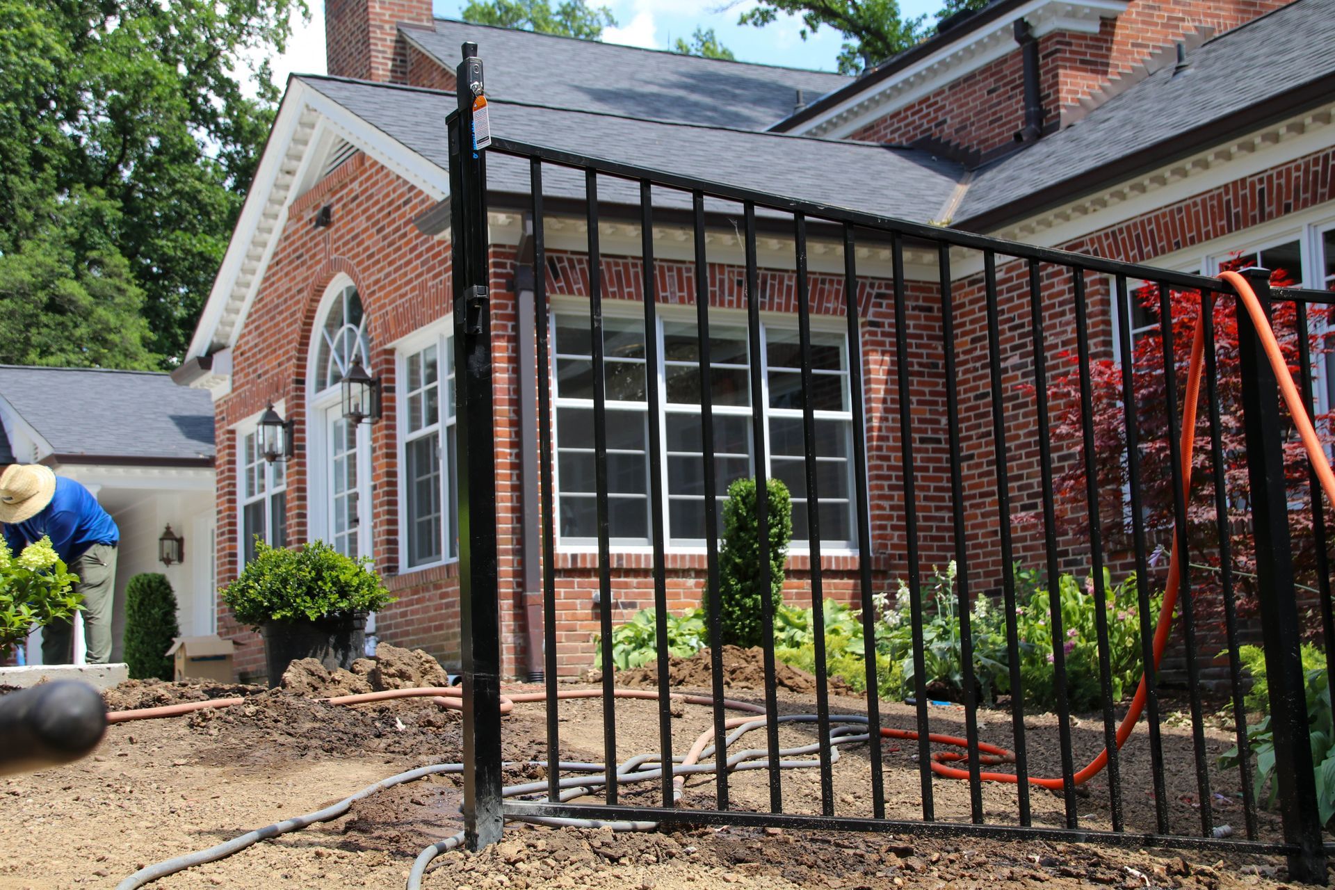 Black metal fence in front of a brick house with a person gardening nearby.