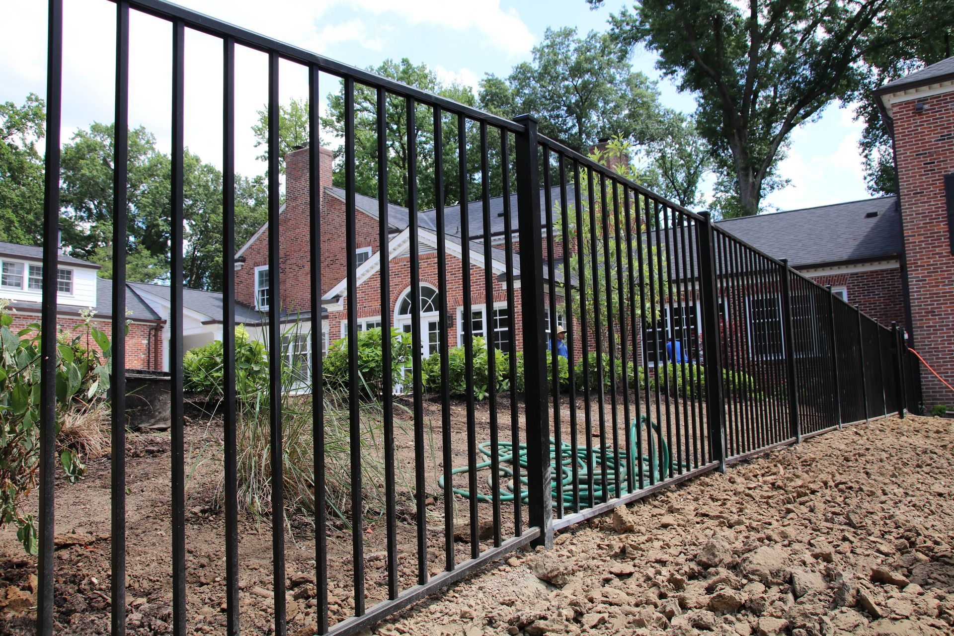 Black metal fence bordering a yard with brown soil, a brick house, and trees in the background.