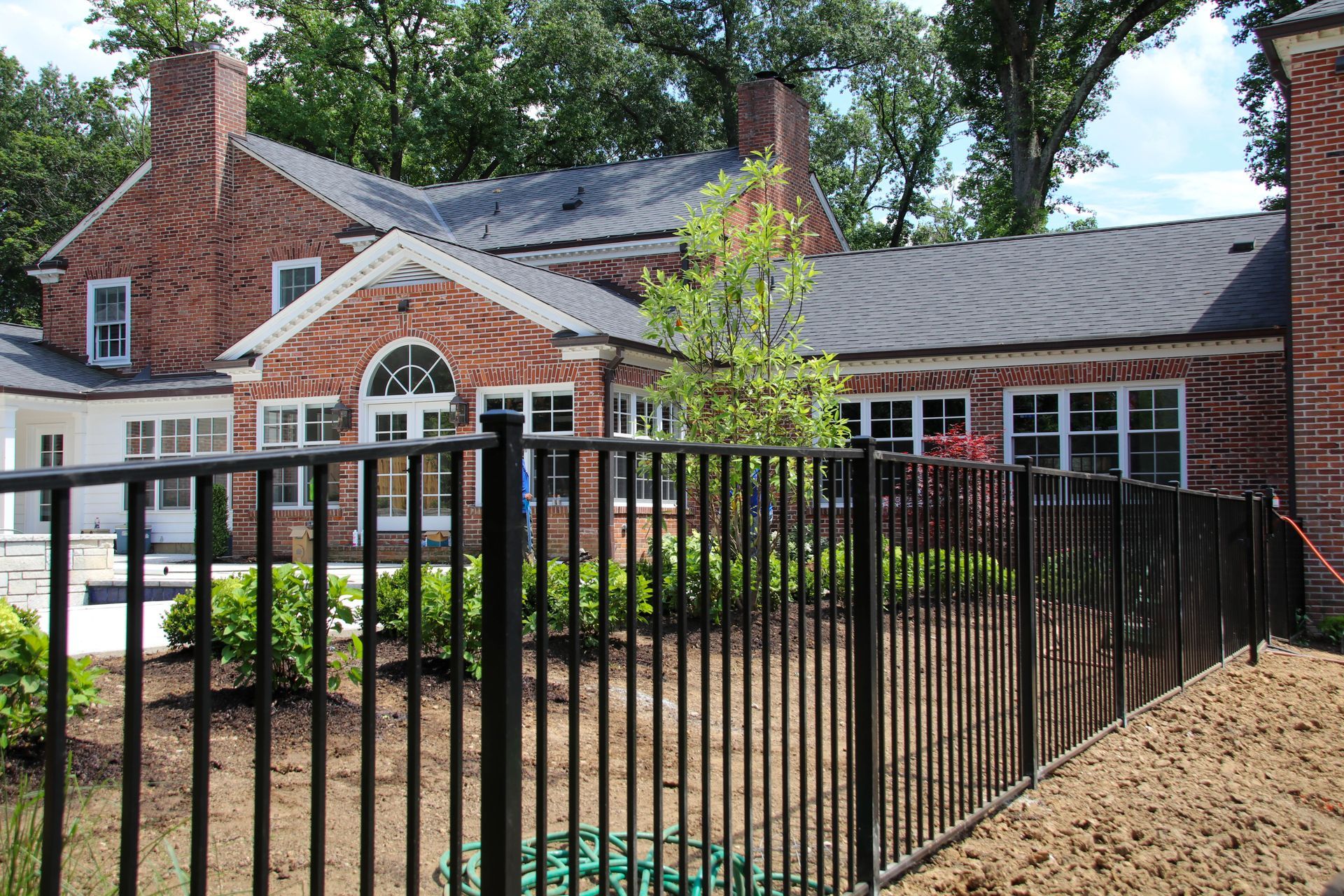 Brick building with a black fence, windows, and a black roof. Green shrubs and trees are in the background.