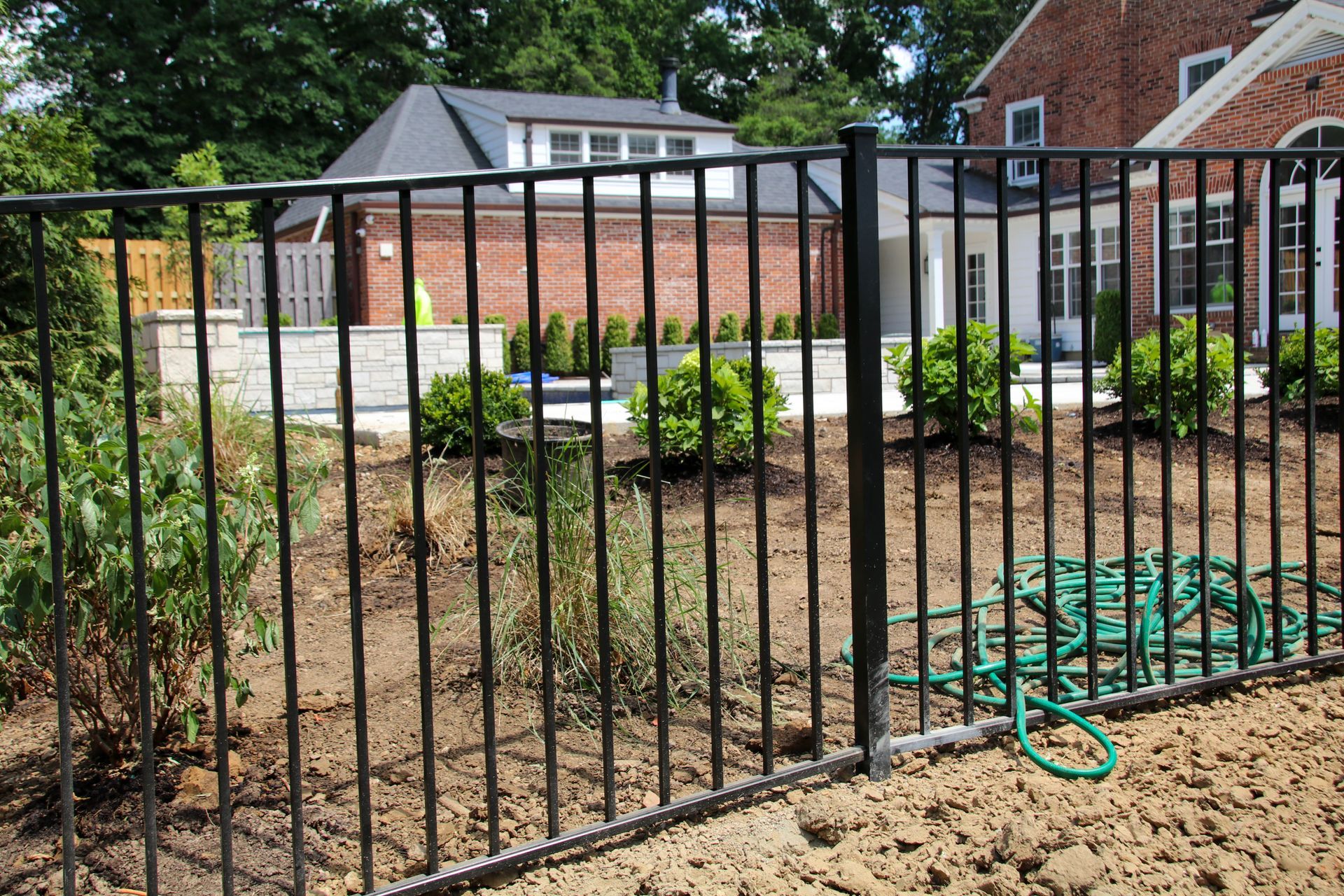 Black metal fence in front of a brick building and backyard with green bushes and a water hose on the ground.