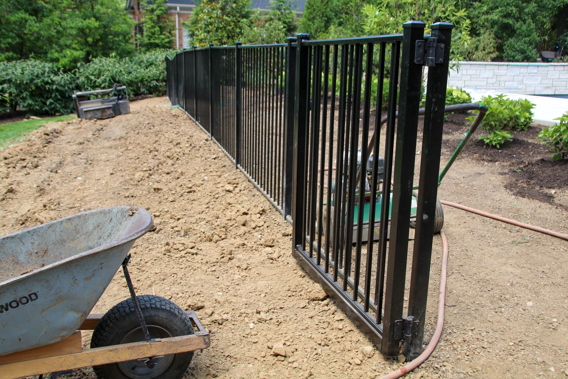 Black metal fence, gate open, being installed in a backyard with a wheelbarrow.