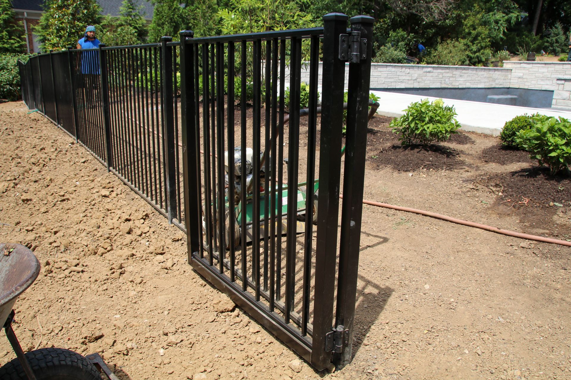 Black metal fence with an open gate in a yard with dirt and a sprinkler.