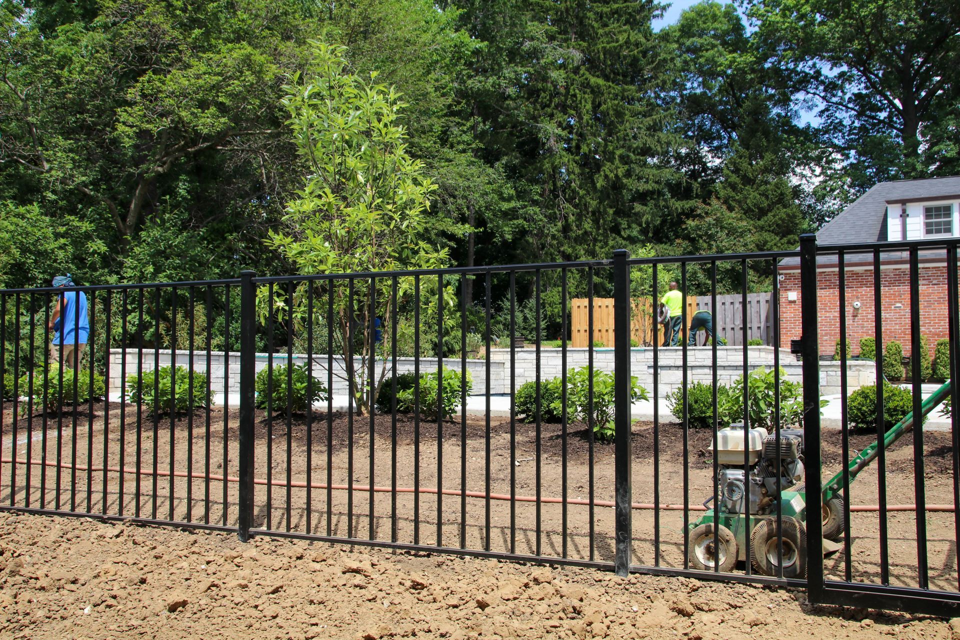 Black metal fence bordering a yard with shrubs, trees, and workers in the background.