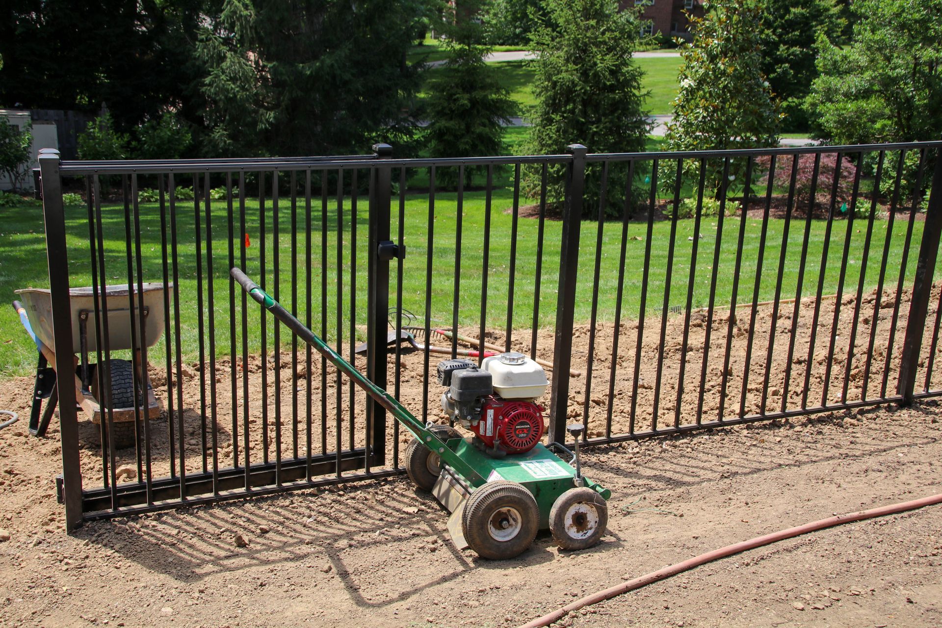 Green aerator near a black metal fence with a gate, on a gravel surface. Trees and lawn in background.