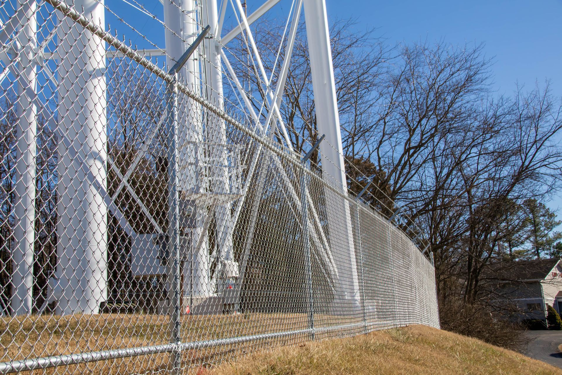 A tall, white water tower behind a chain link fence on a grassy hill; leafless trees in background.